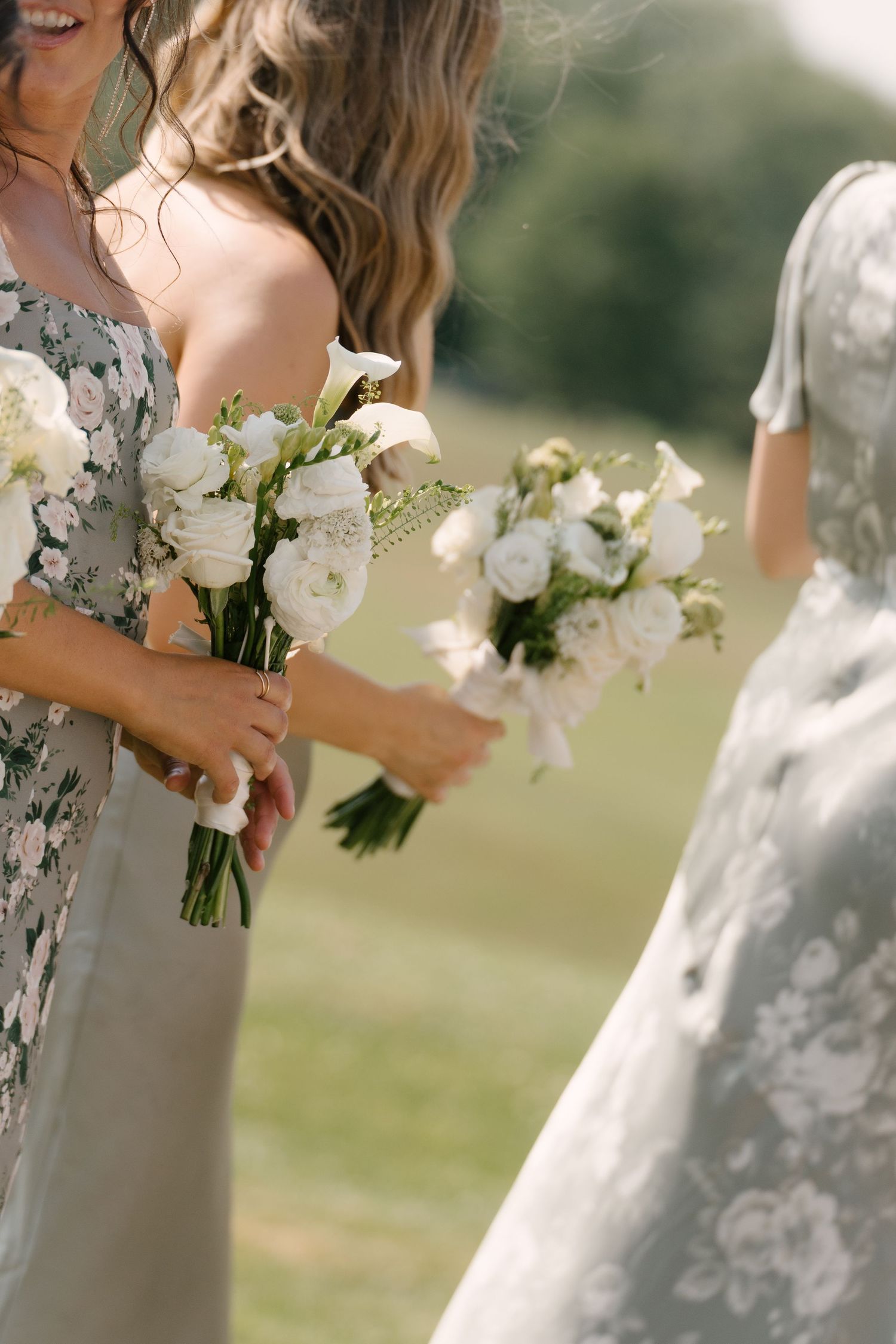 Bridesmaids in floral and sage dresses hold elegant white ranunculus and calla lily bouquets outdoors.