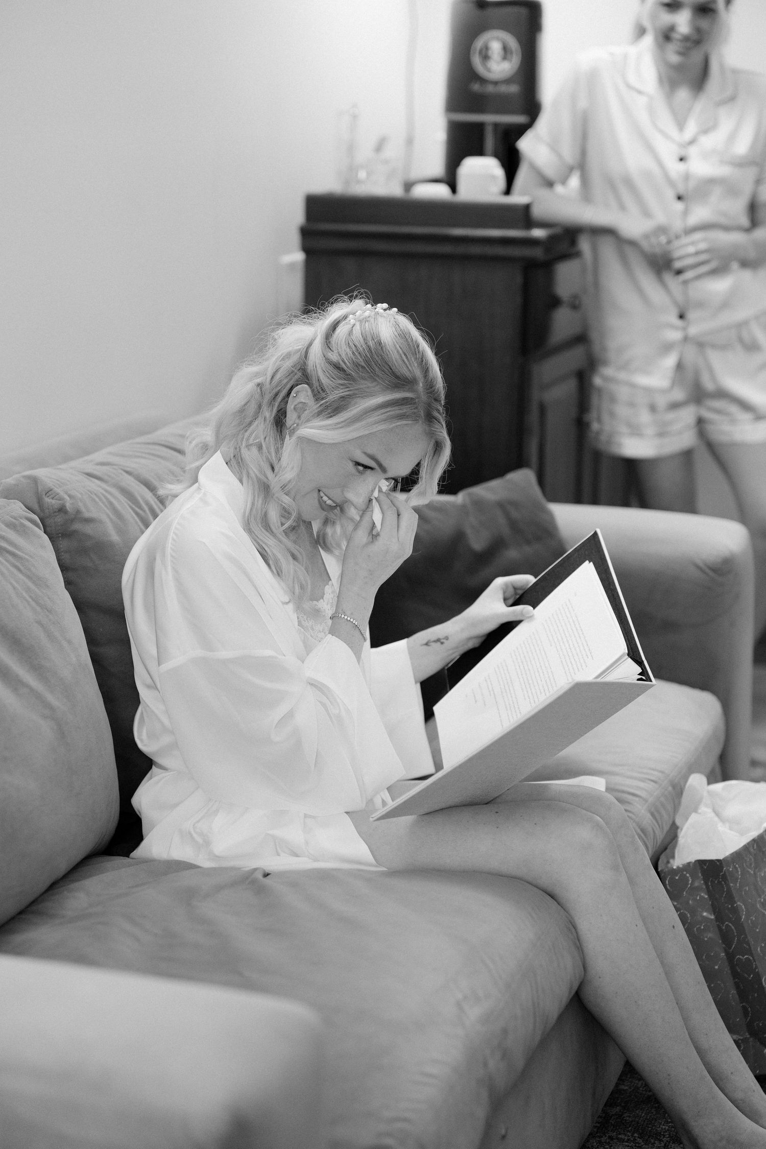Emotional bride in white robe reading heartfelt letter on sofa during wedding morning preparations.