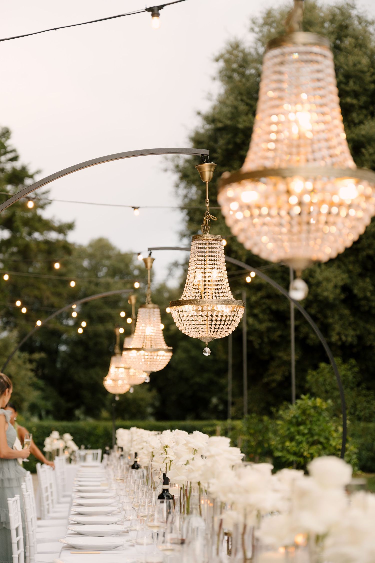 Elegant outdoor Tuscan wedding reception with crystal chandeliers hanging over long table of white roses.
