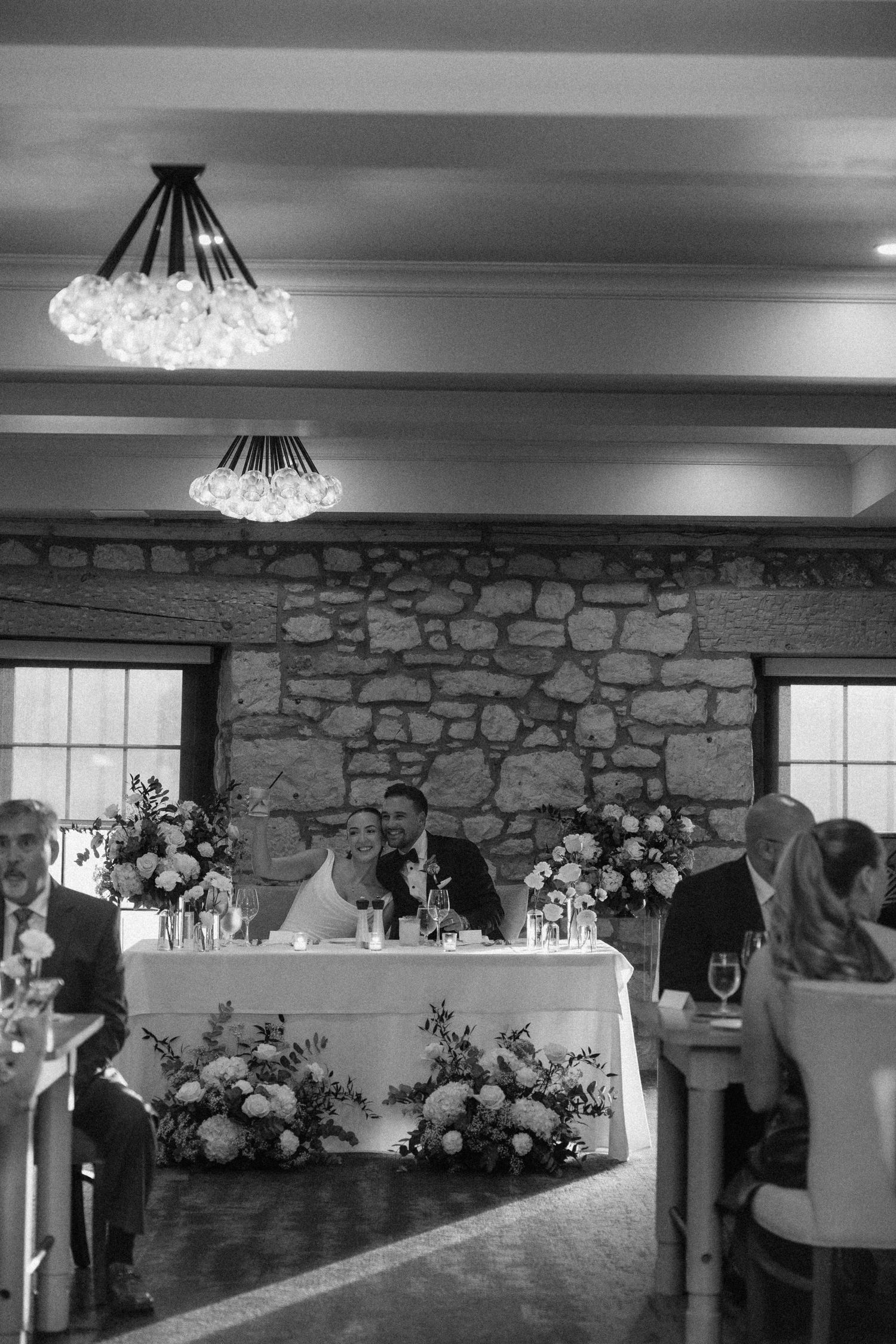 Bride and groom seated at sweetheart table adorned with floral arrangements in stone-walled reception hall at Cambridge Mill.