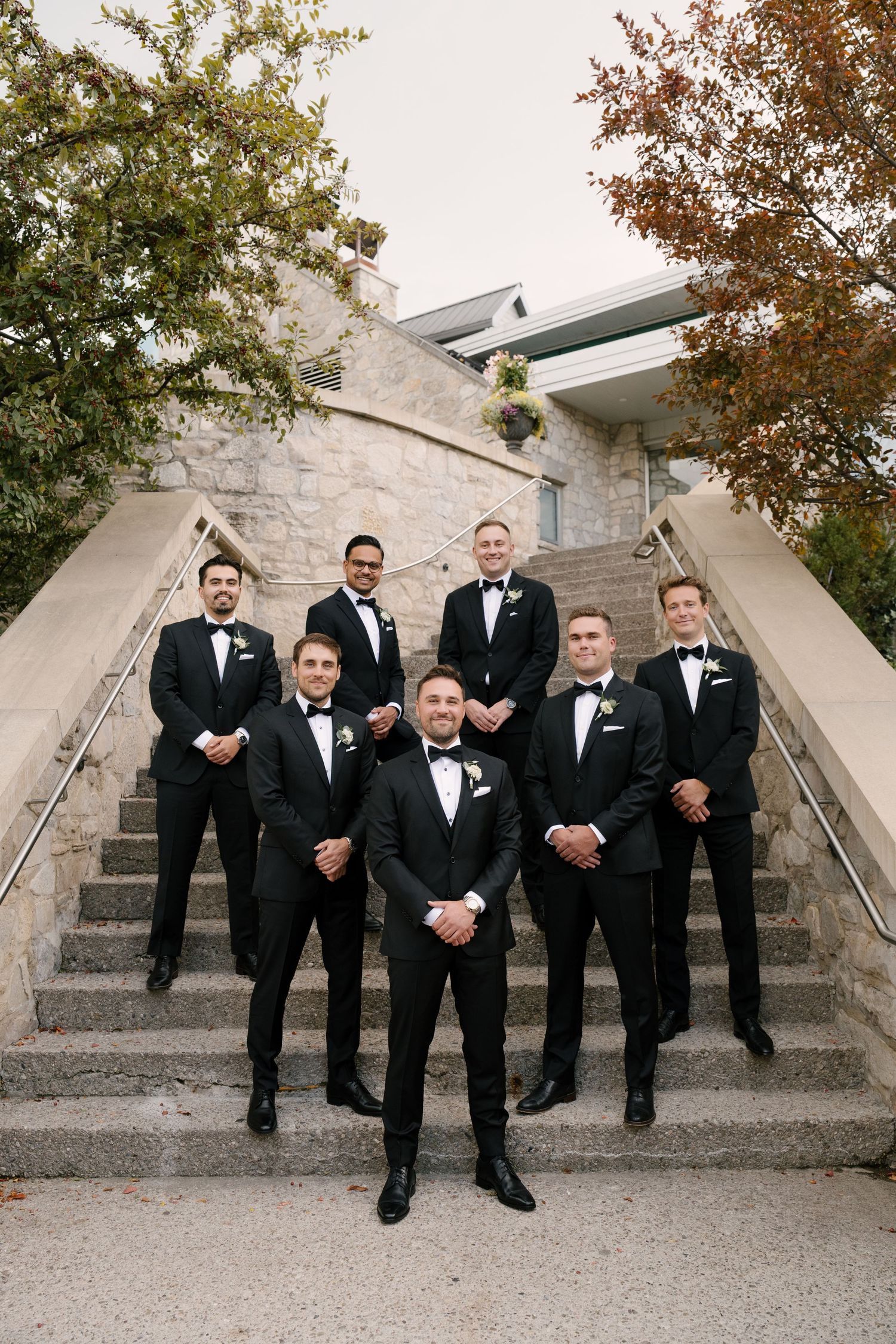 Groom and groomsmen in black tuxedos with bow ties pose on stone steps outside Cambridge Mill for a wedding photo.