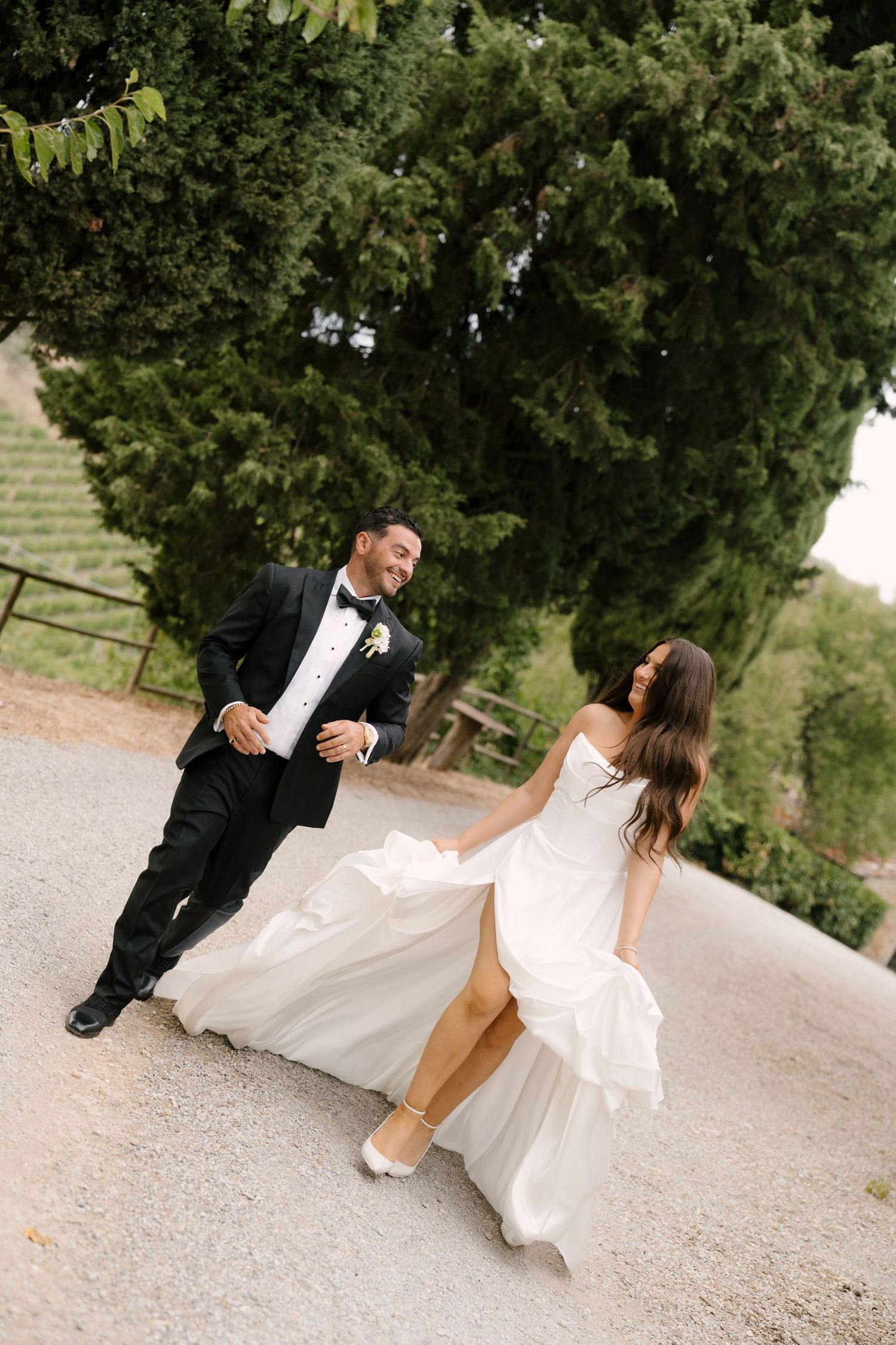 Joyful bride and groom in formal attire walk hand-in-hand on gravel path surrounded by cypress trees at Tuscan Villa.
