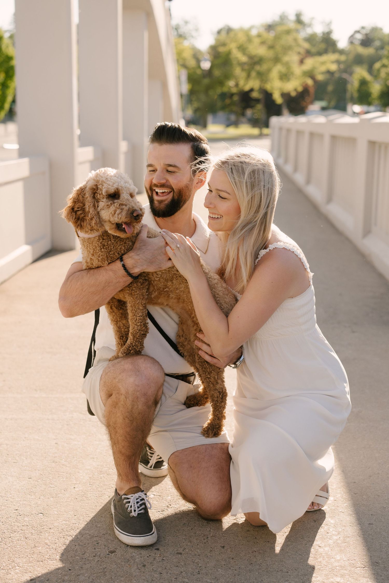 Smiling couple crouching with fluffy brown Goldendoodle on bridge.