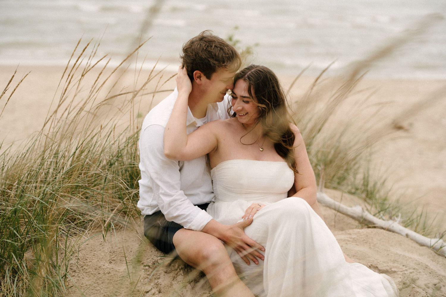 Couple sits together in beach sand dunes, smiling intimately as tall grass sways around them.