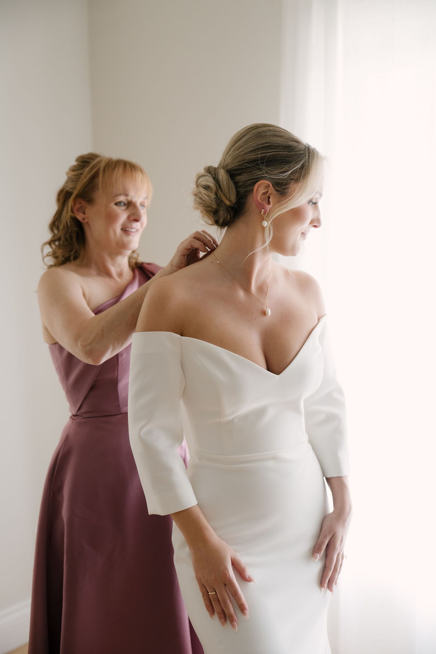 Bride in white off-shoulder gown gets jewelry help from bridesmaid in mauve dress near bright window.