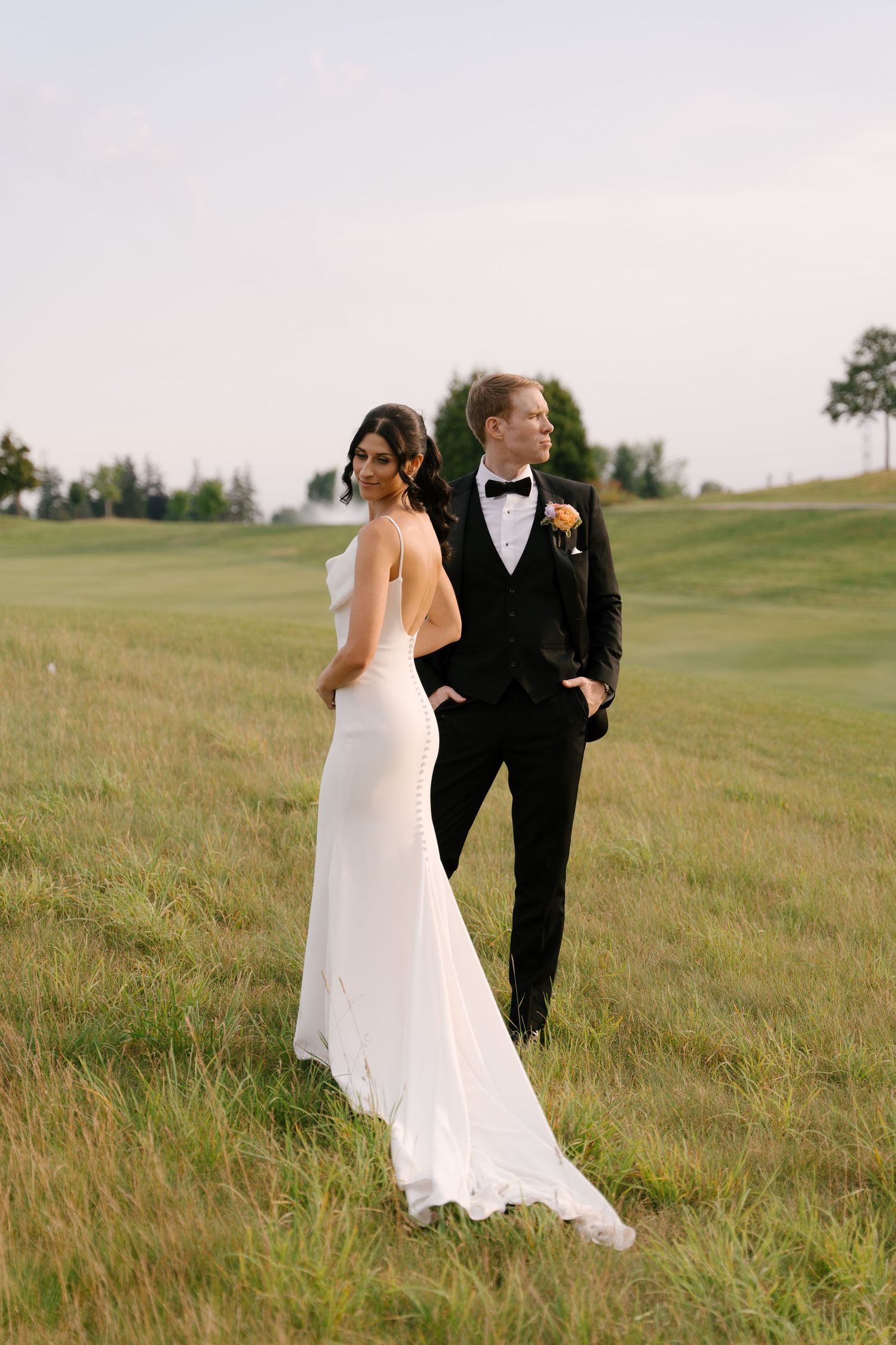 Bride in white gown and groom in black tuxedo standing in lush green field at dusk at Whistle Bear Venue.