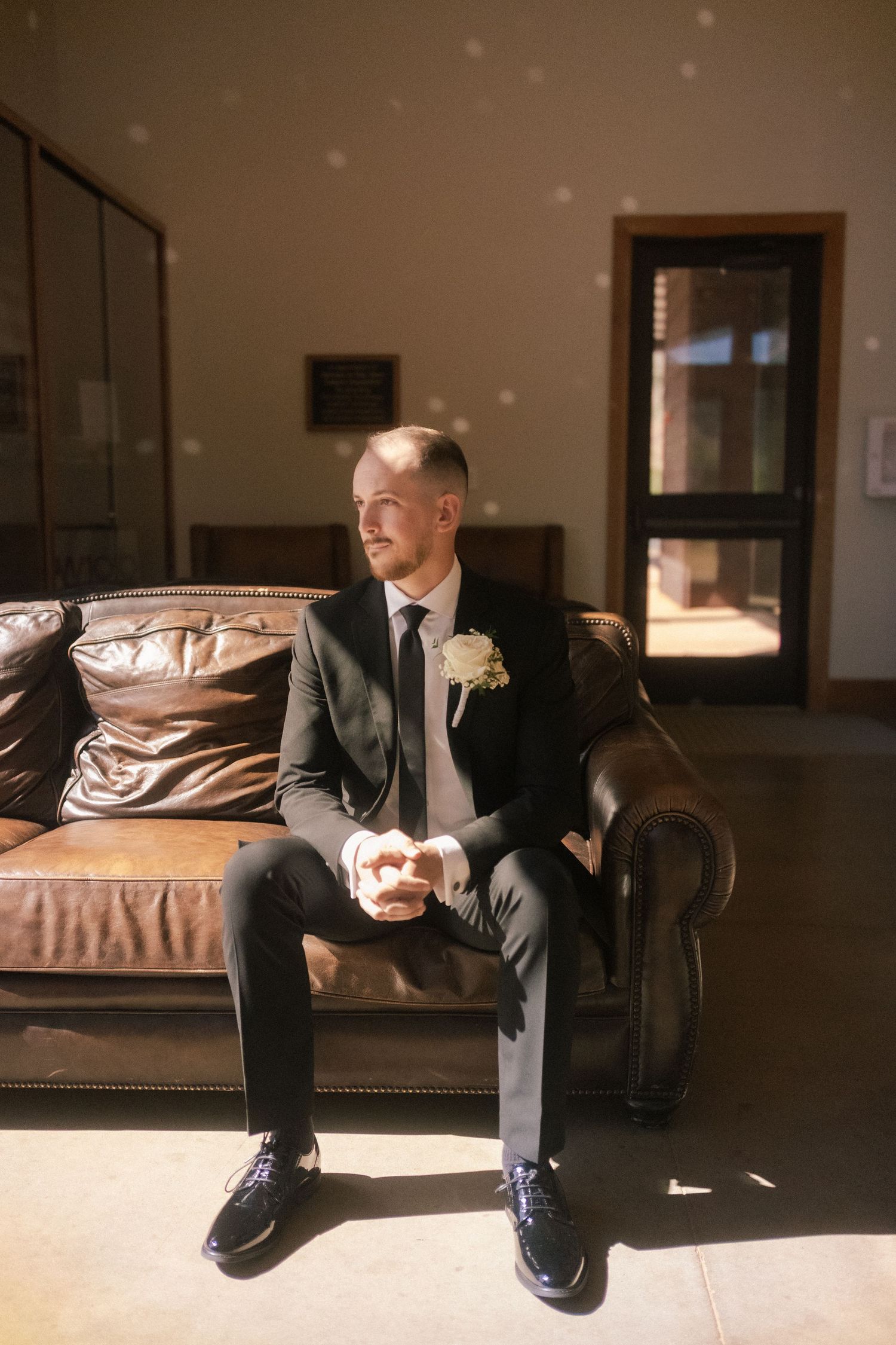 Groom in classic black suit sitting on leather couch with boutonniere.