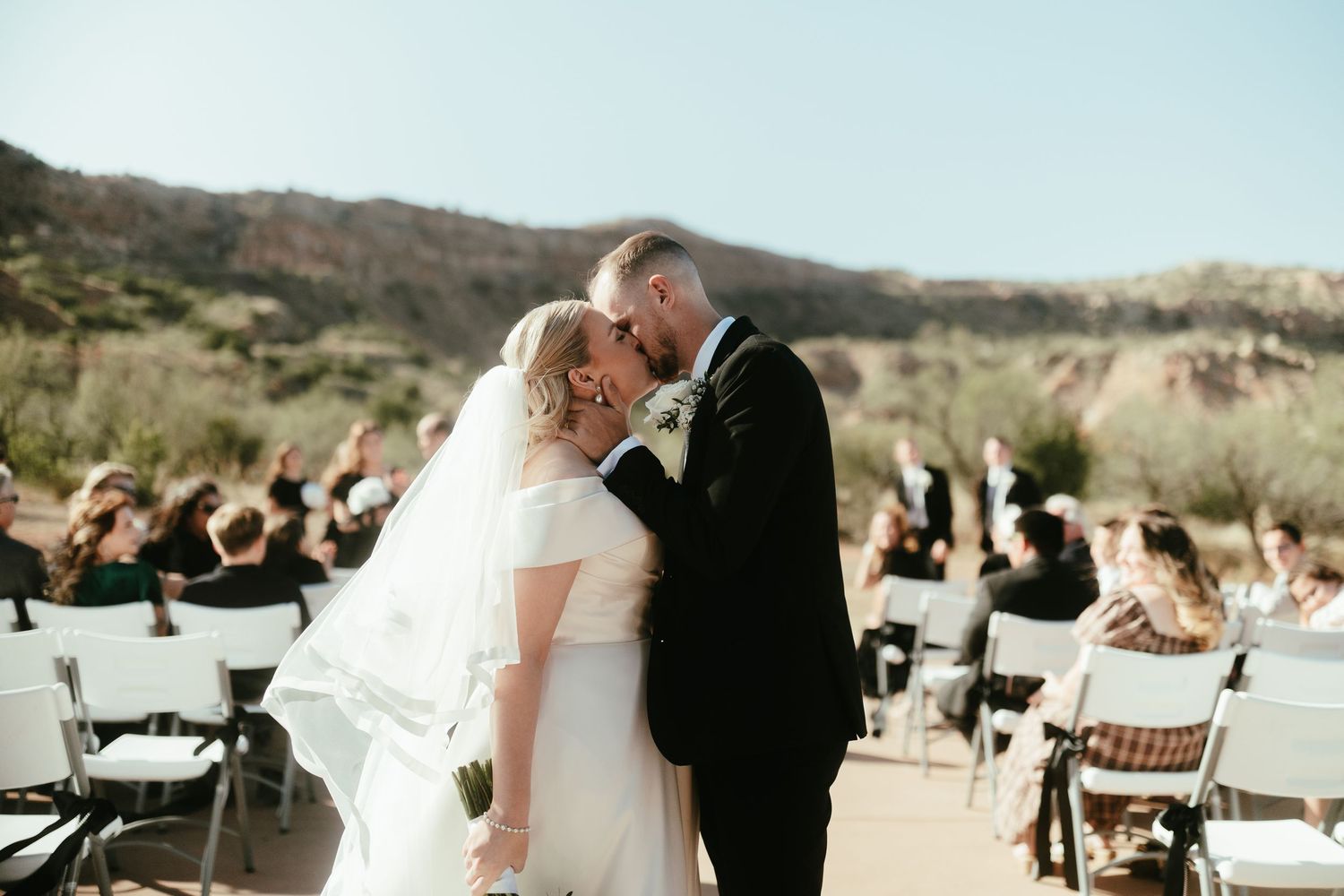 Outdoor wedding ceremony setup with chairs and guests in natural desert landscape.