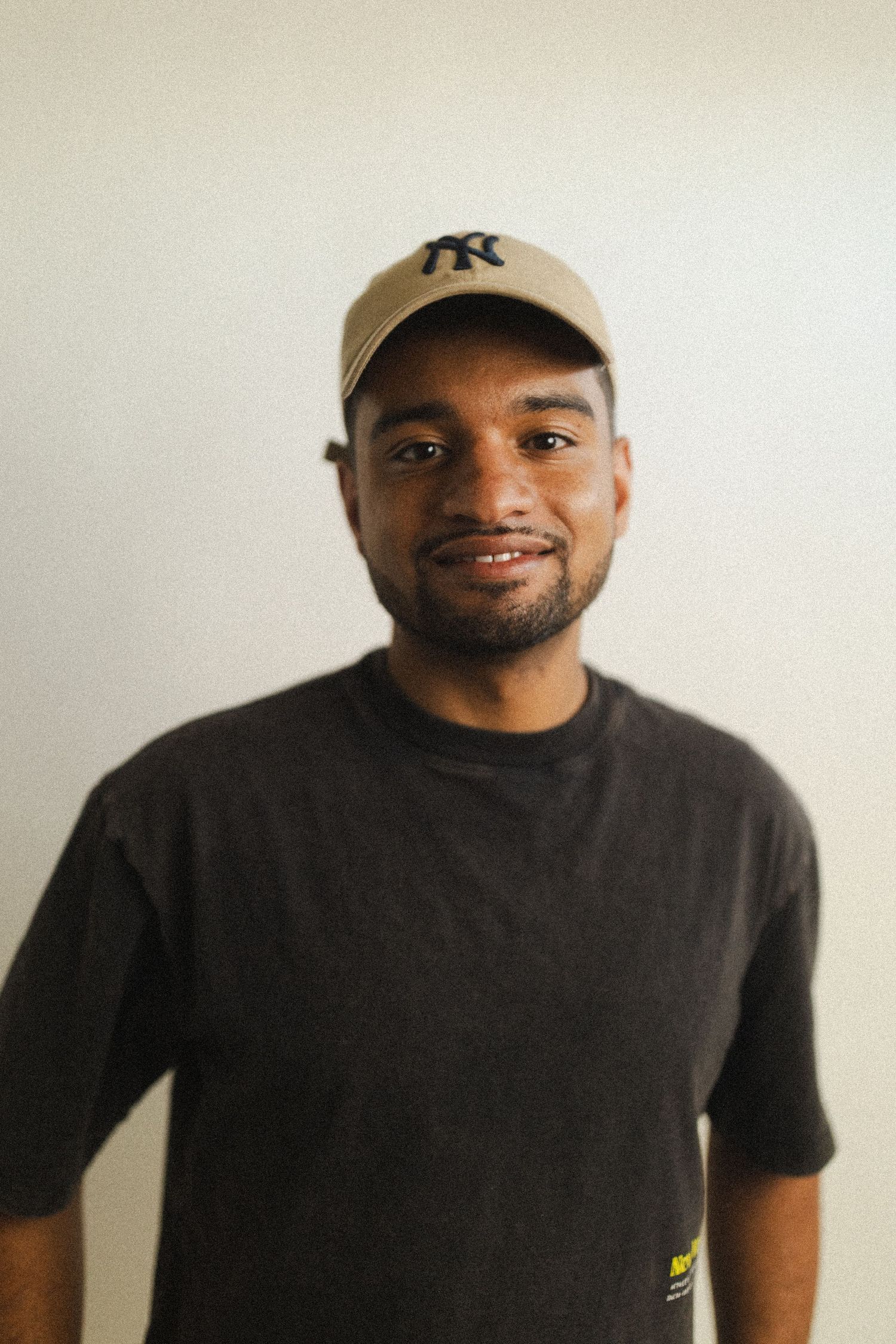 Young man smiling, wearing a tan NY Yankees cap and black t-shirt, standing against a white wall.