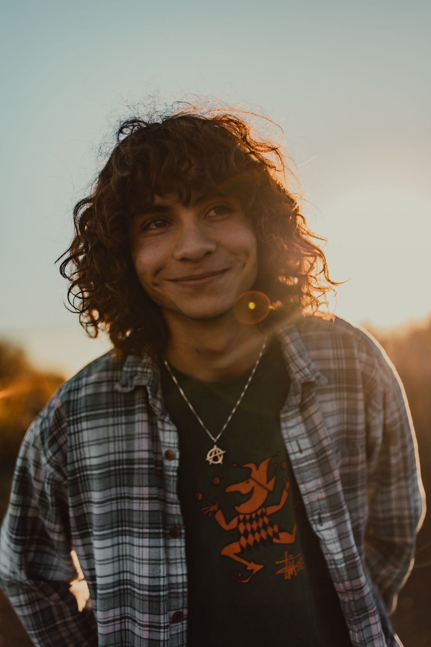 Candid portrait of someone with curly hair smiling in plaid shirt at sunset.