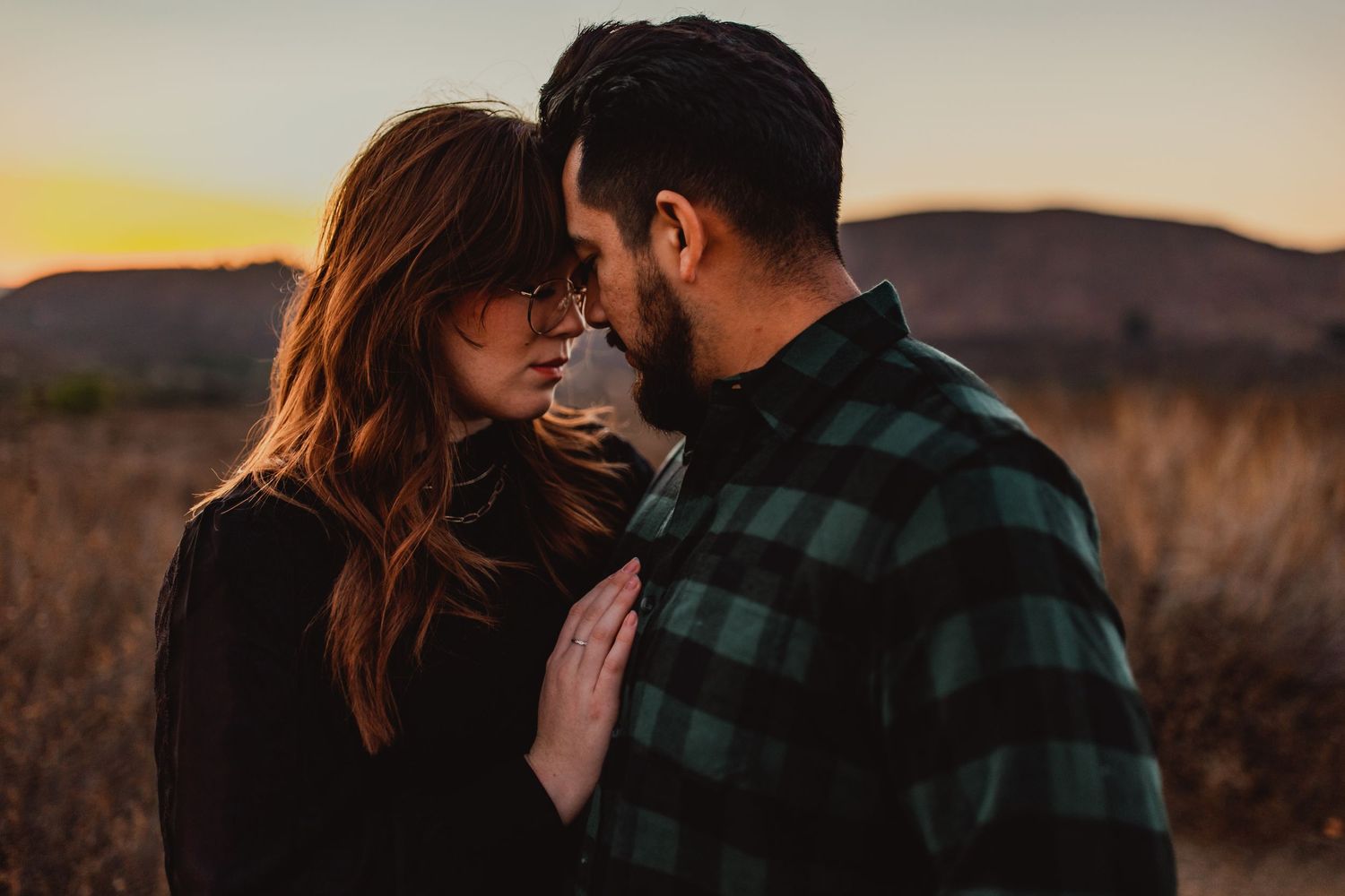 Romantic sunset photos of a couple embracing in a field wearing casual plaid clothing.