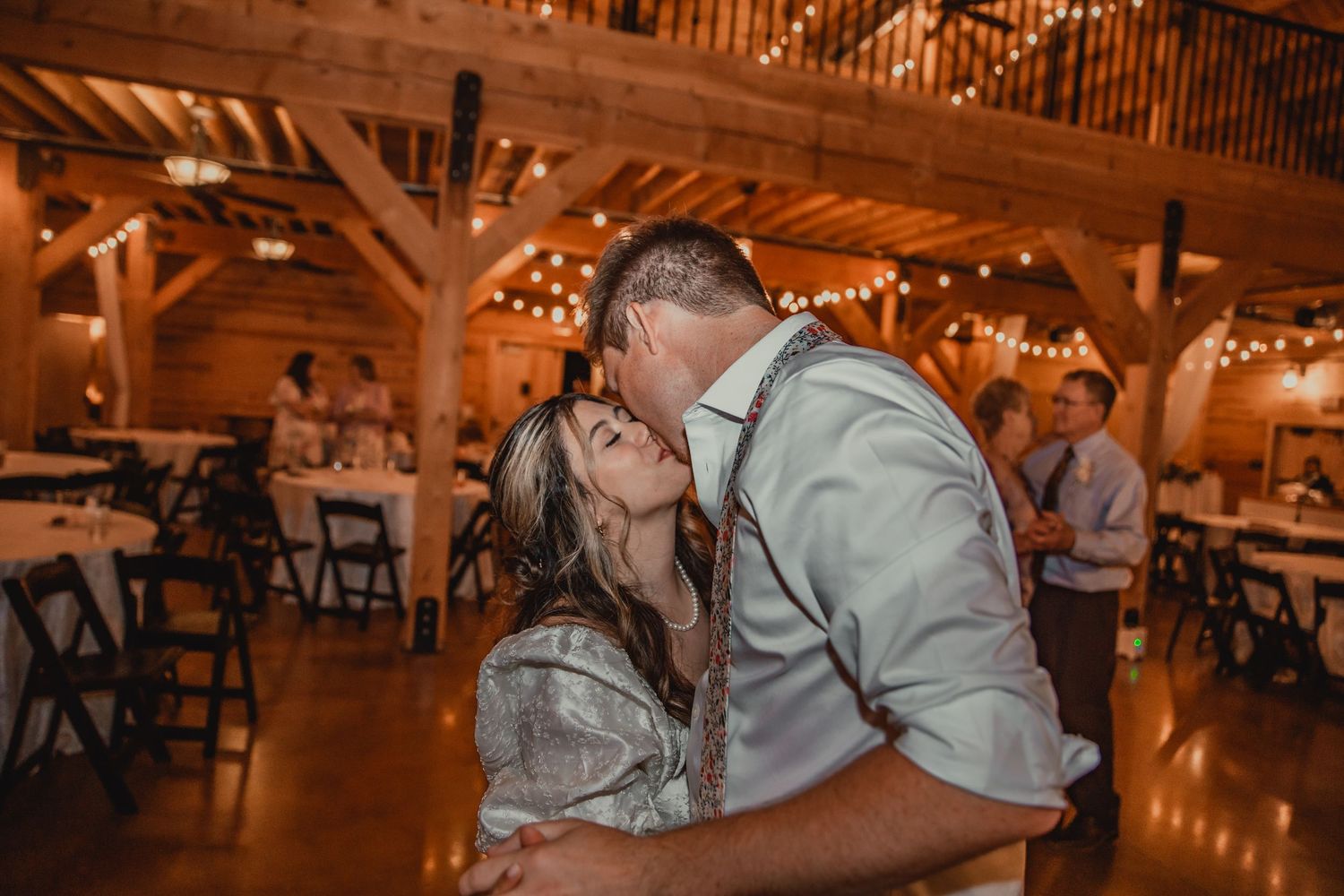 Couple sharing romantic moment in rustic barn venue with string lights overhead.