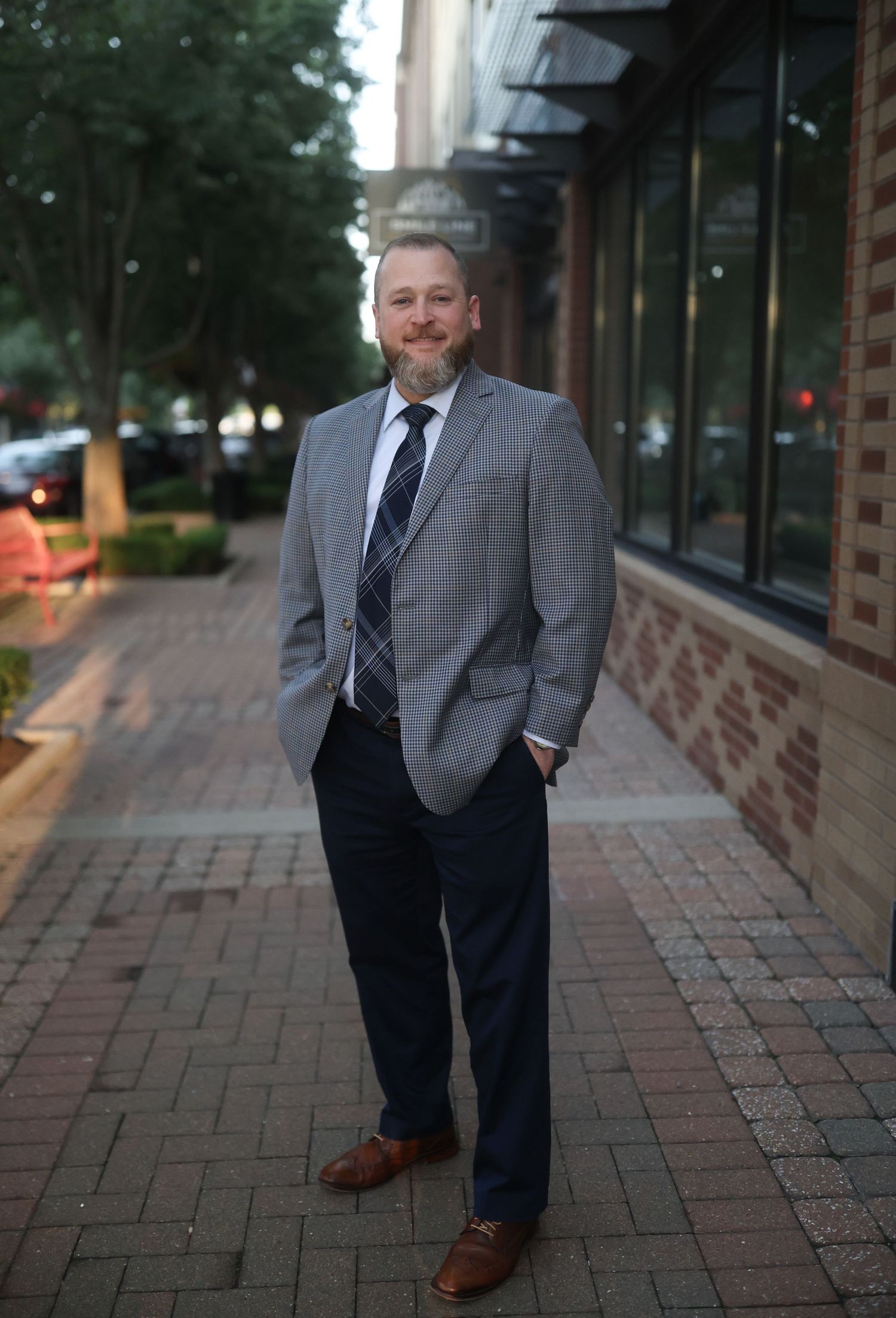 Man in formal business attire poses on brick sidewalk with building exterior in background.