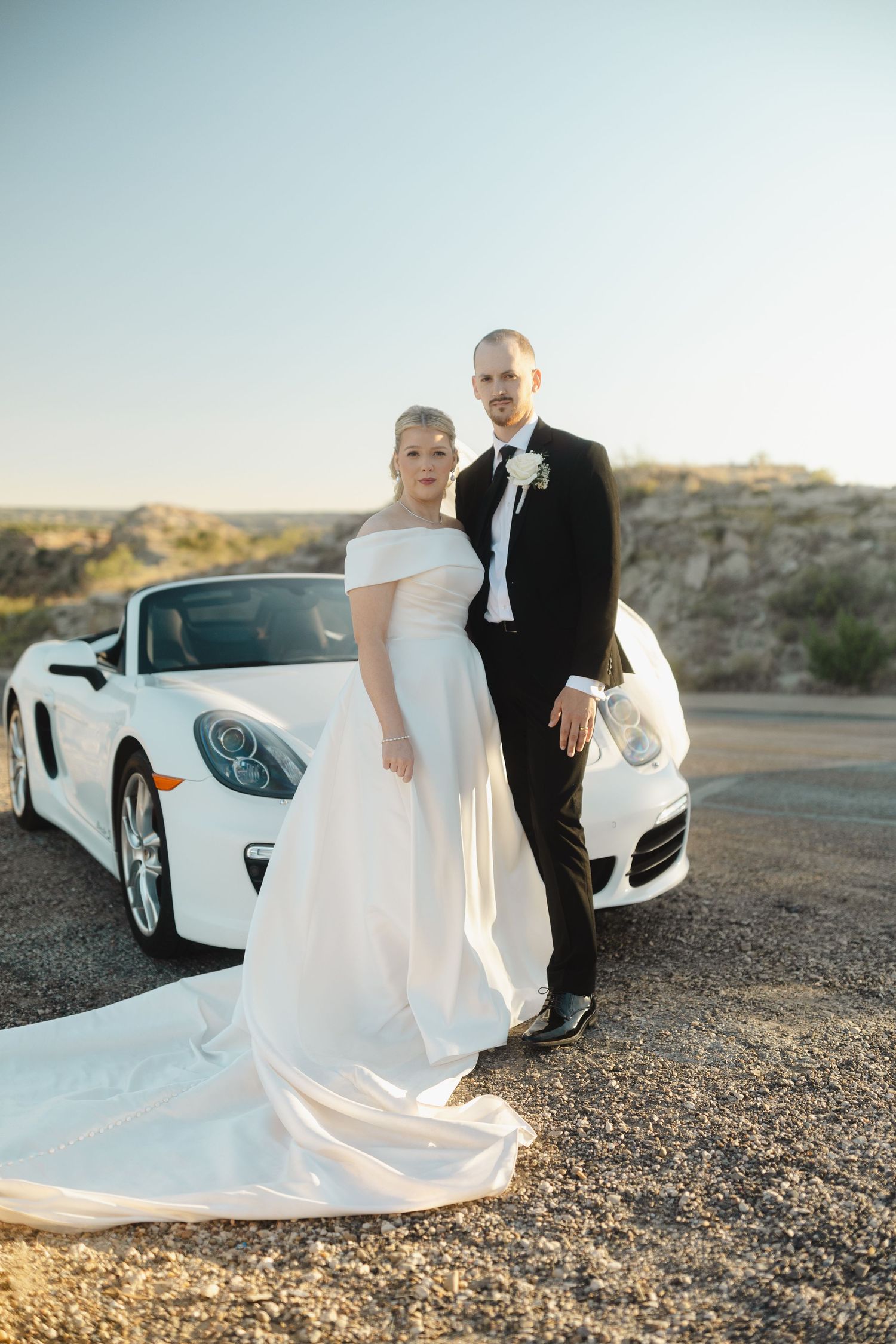 A bride and groom pose elegantly next to a white luxury sports car in a desert setting.