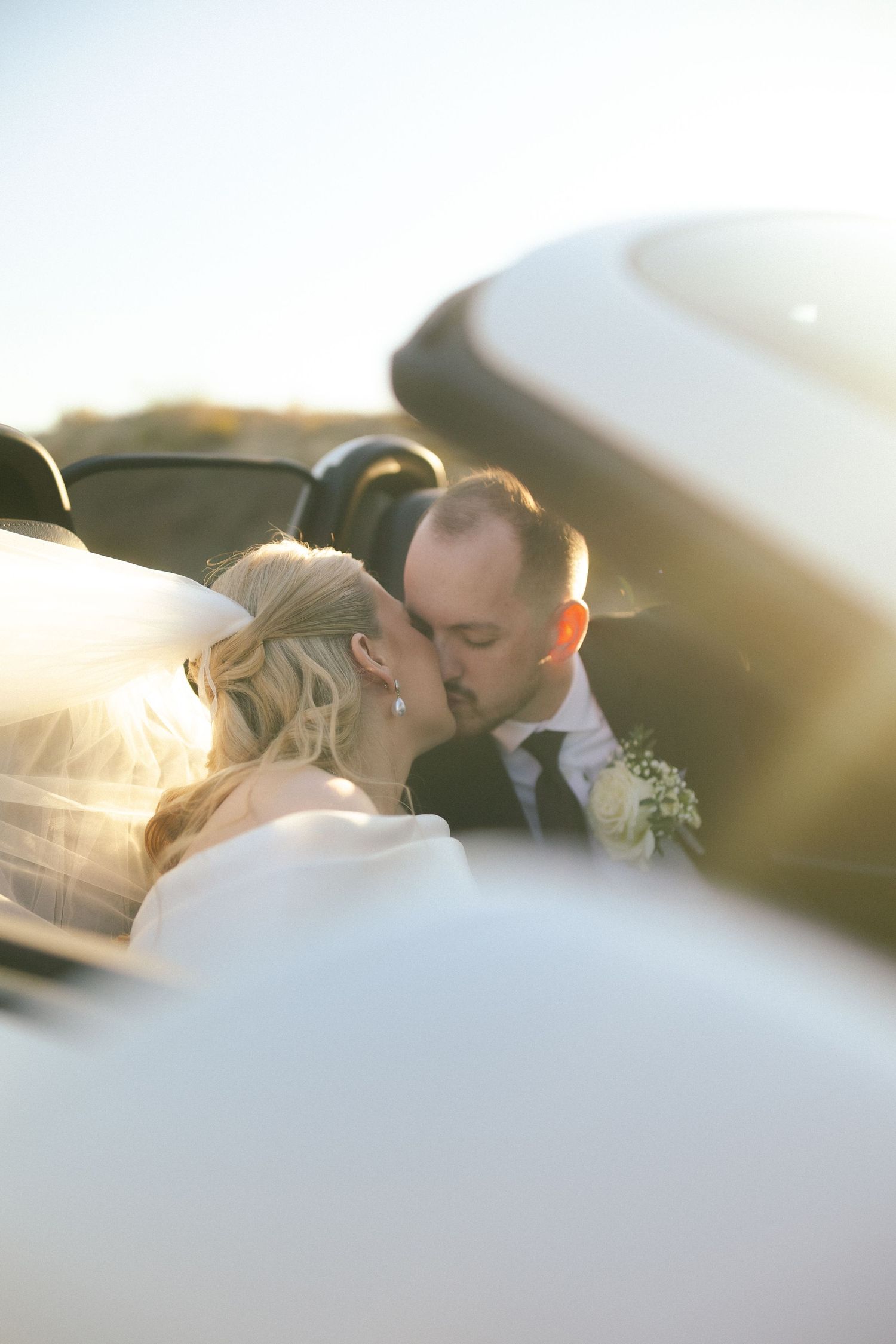Romantic wedding kiss captured through car window at sunset.