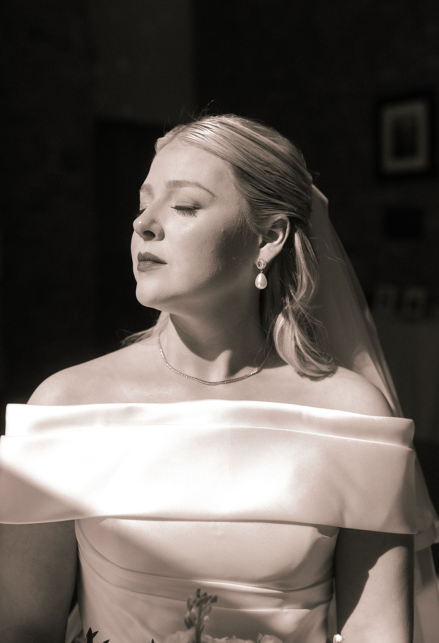 Elegant bride in off-shoulder wedding gown with pearl earrings in dramatic lighting.