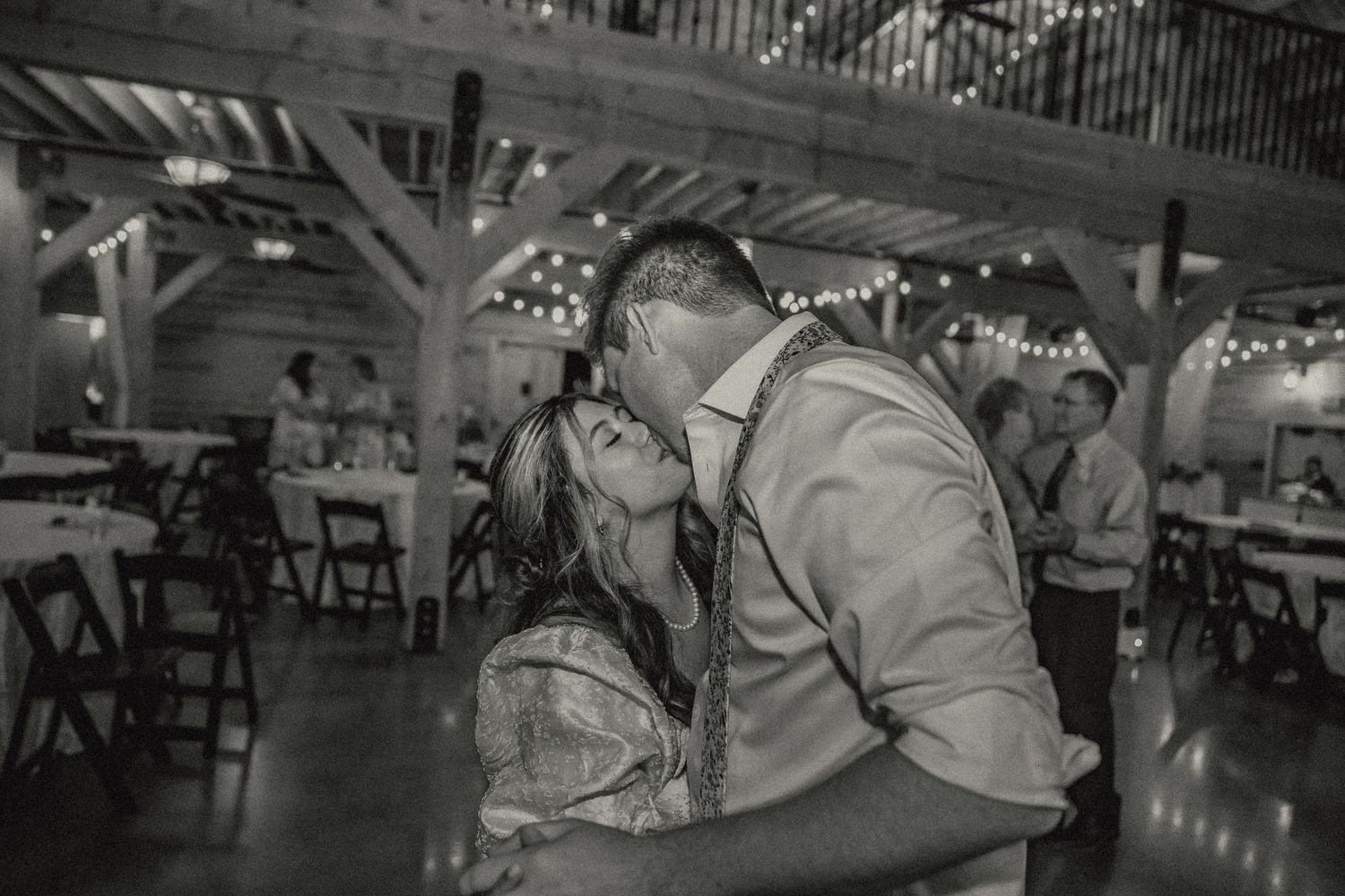 Romantic couple sharing an intimate dance moment under twinkling lights at a rustic reception venue.