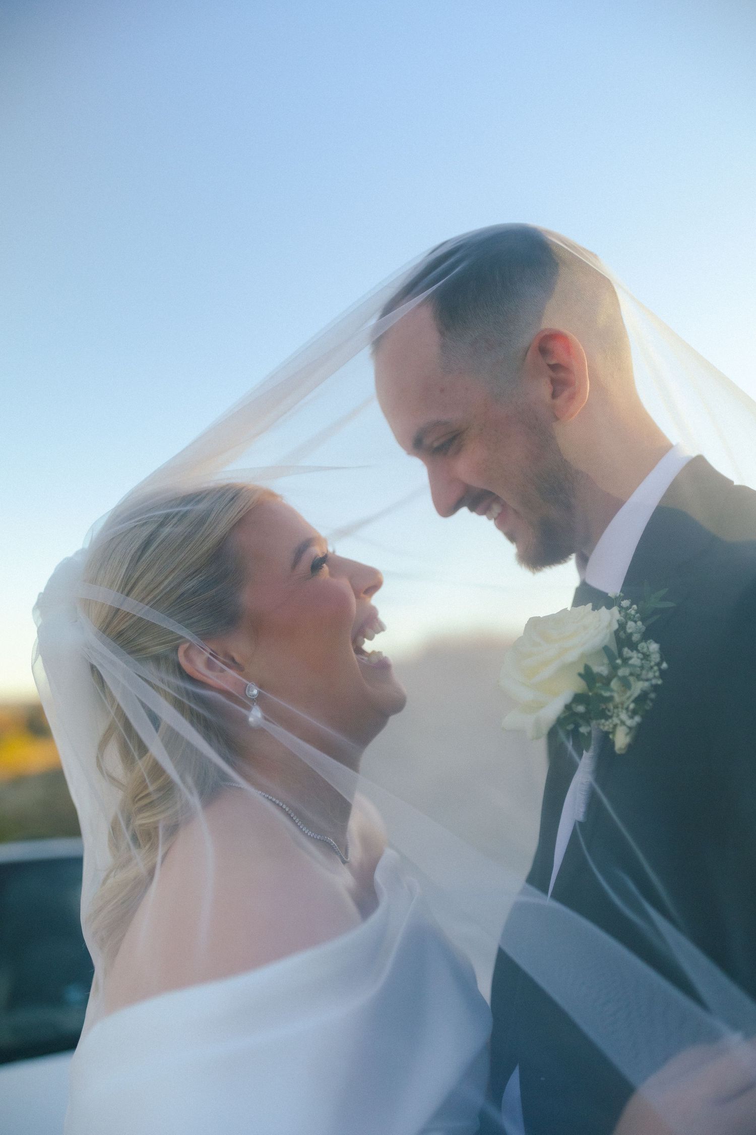 Romantic outdoor wedding portrait with couple embracing under a flowing veil against blue sky.