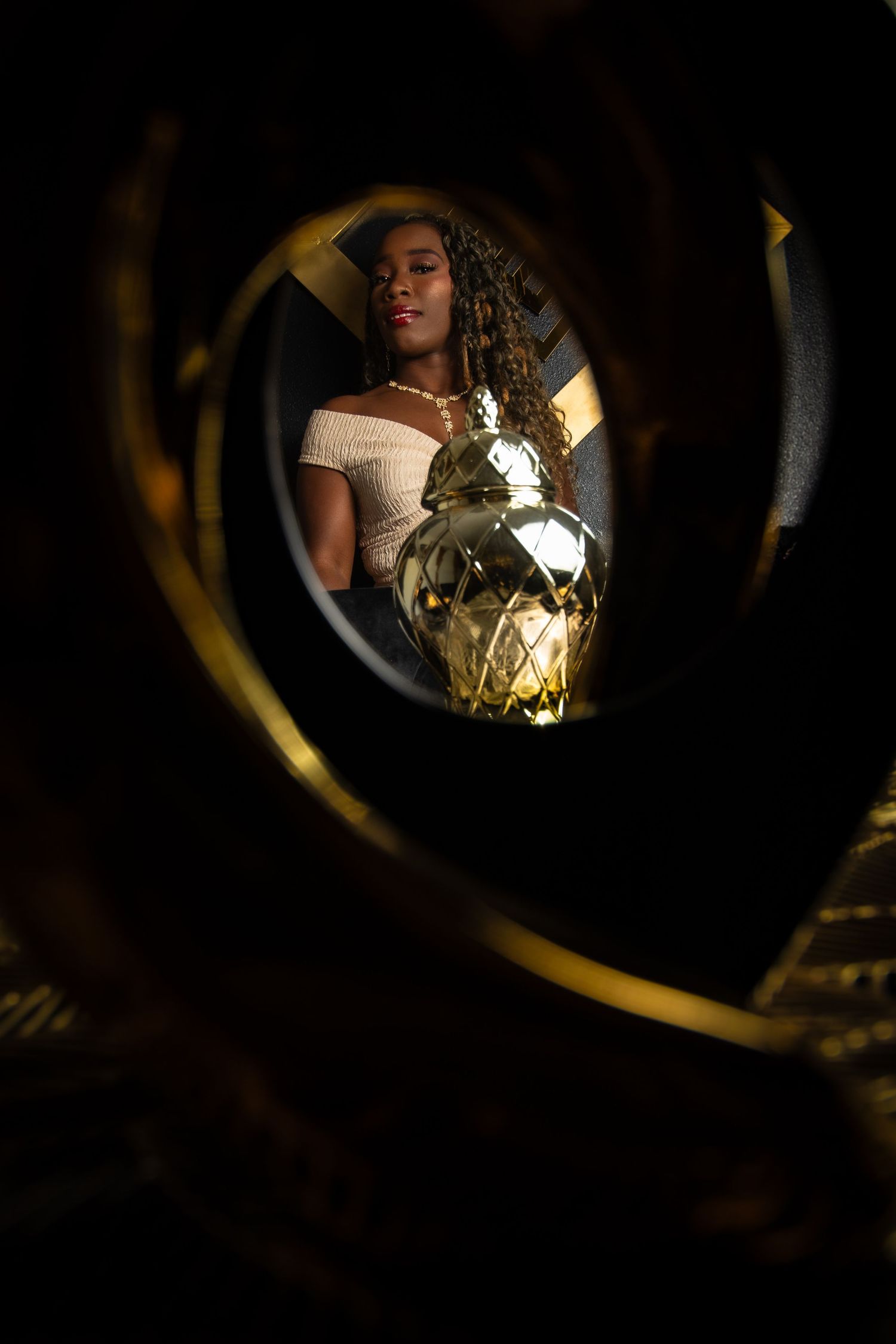 Young woman with braids holding a gold disco ball, viewed through a circular frame in dramatic dark lighting.
