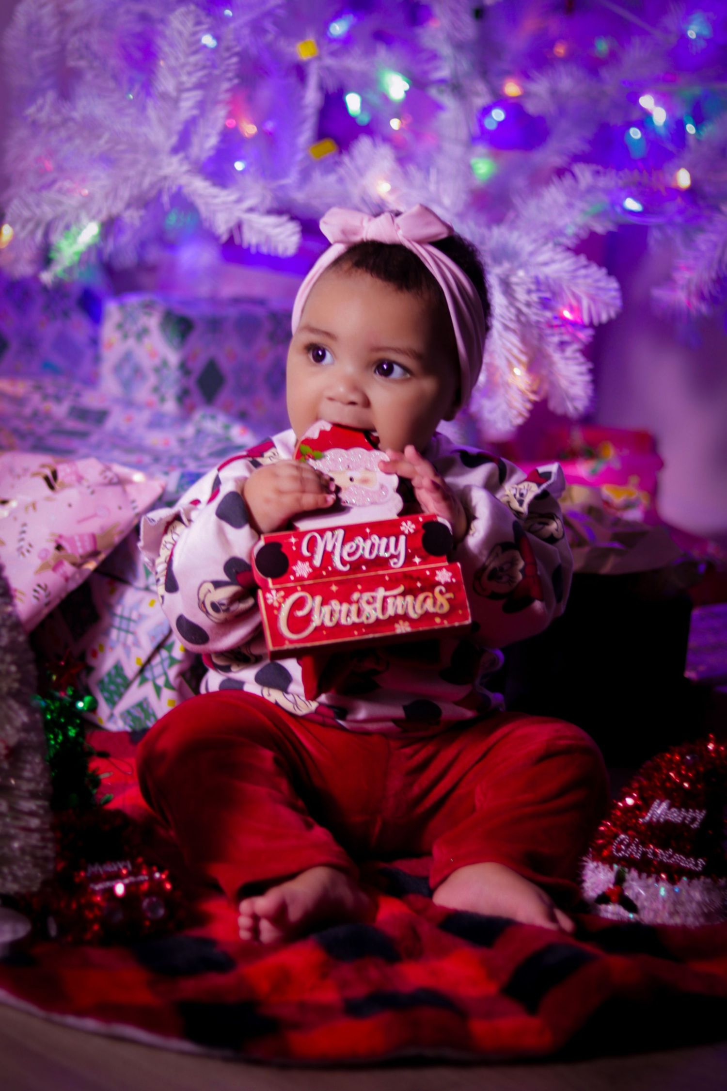 Baby in red holiday outfit poses near purple-lit Christmas tree with festive decorations.