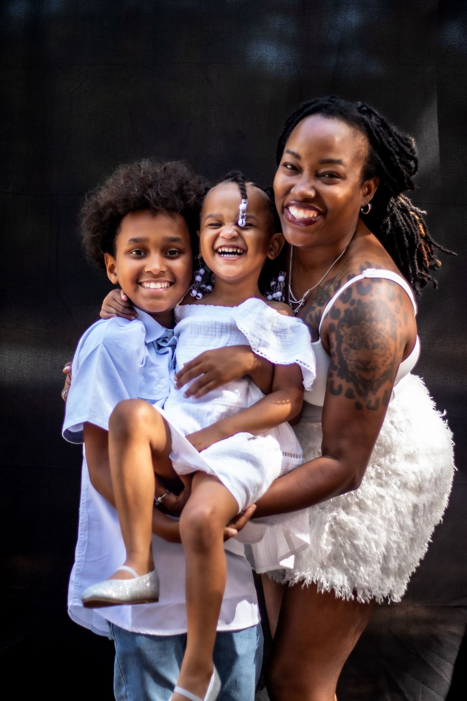 A smiling Black mother with tattoos and locs holds two young children dressed in white outdoors.