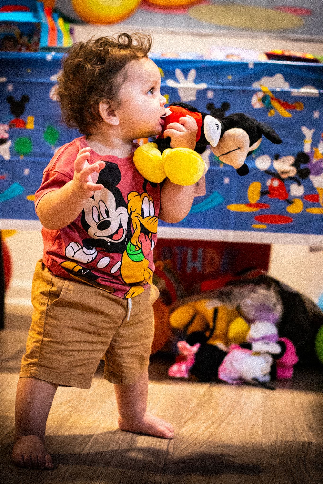 Toddler in brown shorts plays with Disney plush toys against decorated wall.