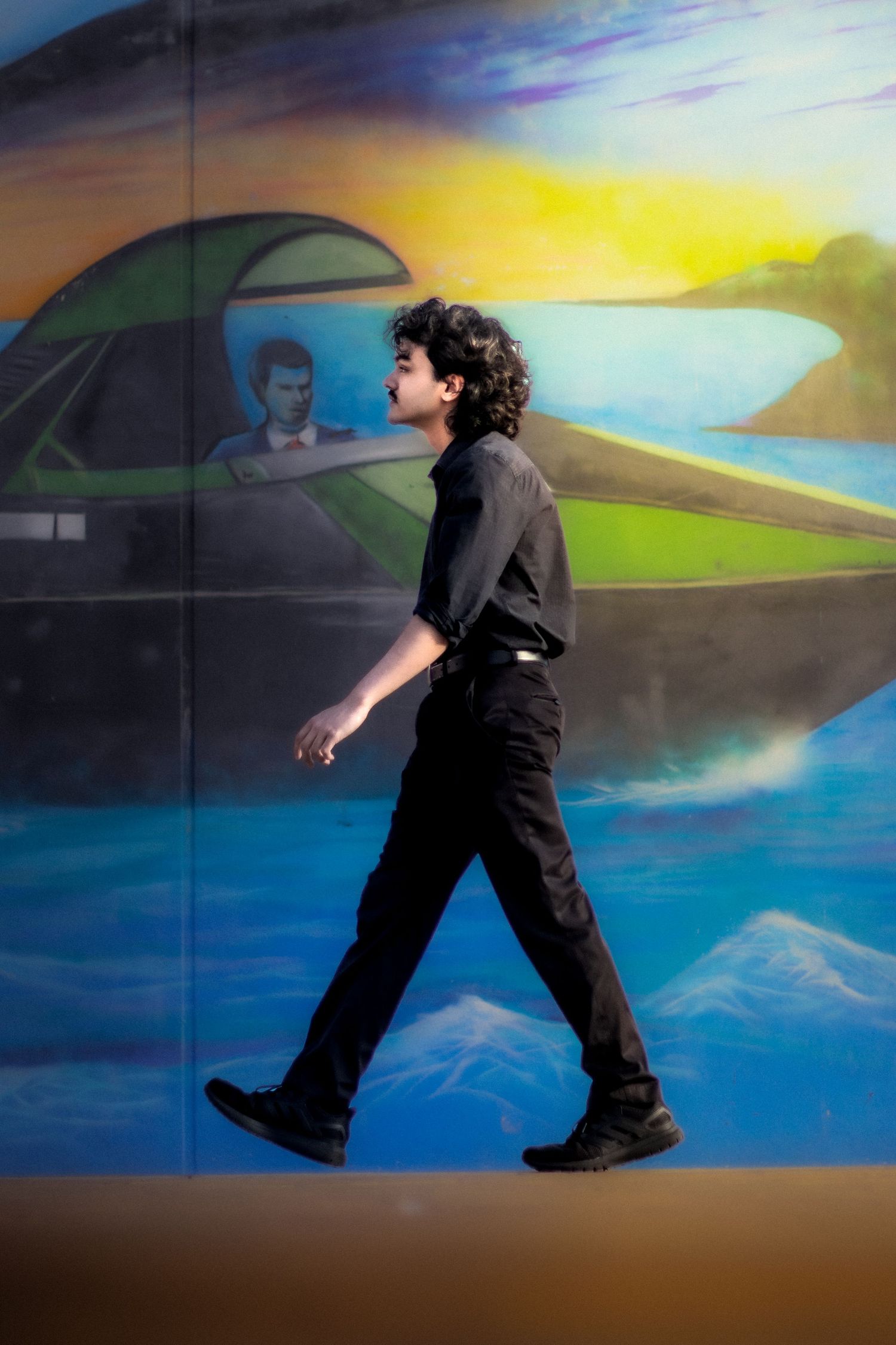Young man in black outfit walking past a vibrant colorful mural depicting a boat on water with mountains.