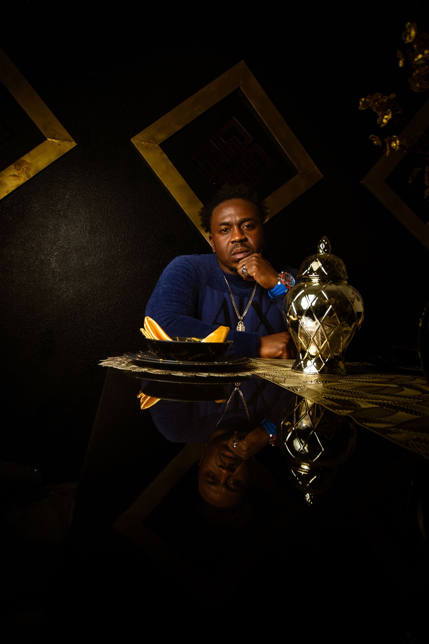 Man in blue shirt sitting at reflective table with trophy in dark room with gold diamond wall decor.