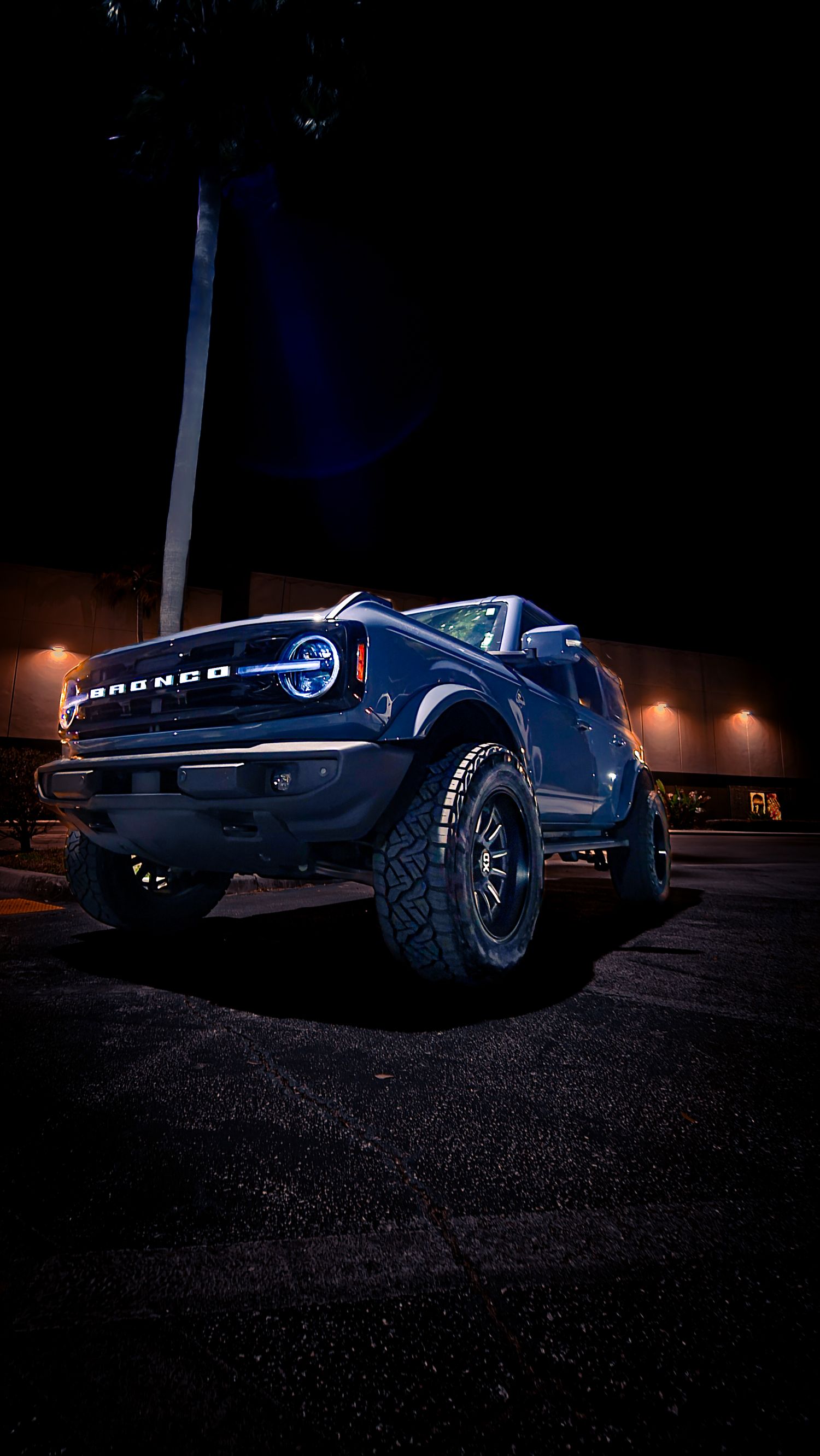 A lifted Ford Bronco SUV with glowing LED headlights parked in a dark lot at night, showcasing its rugged off-road tires.