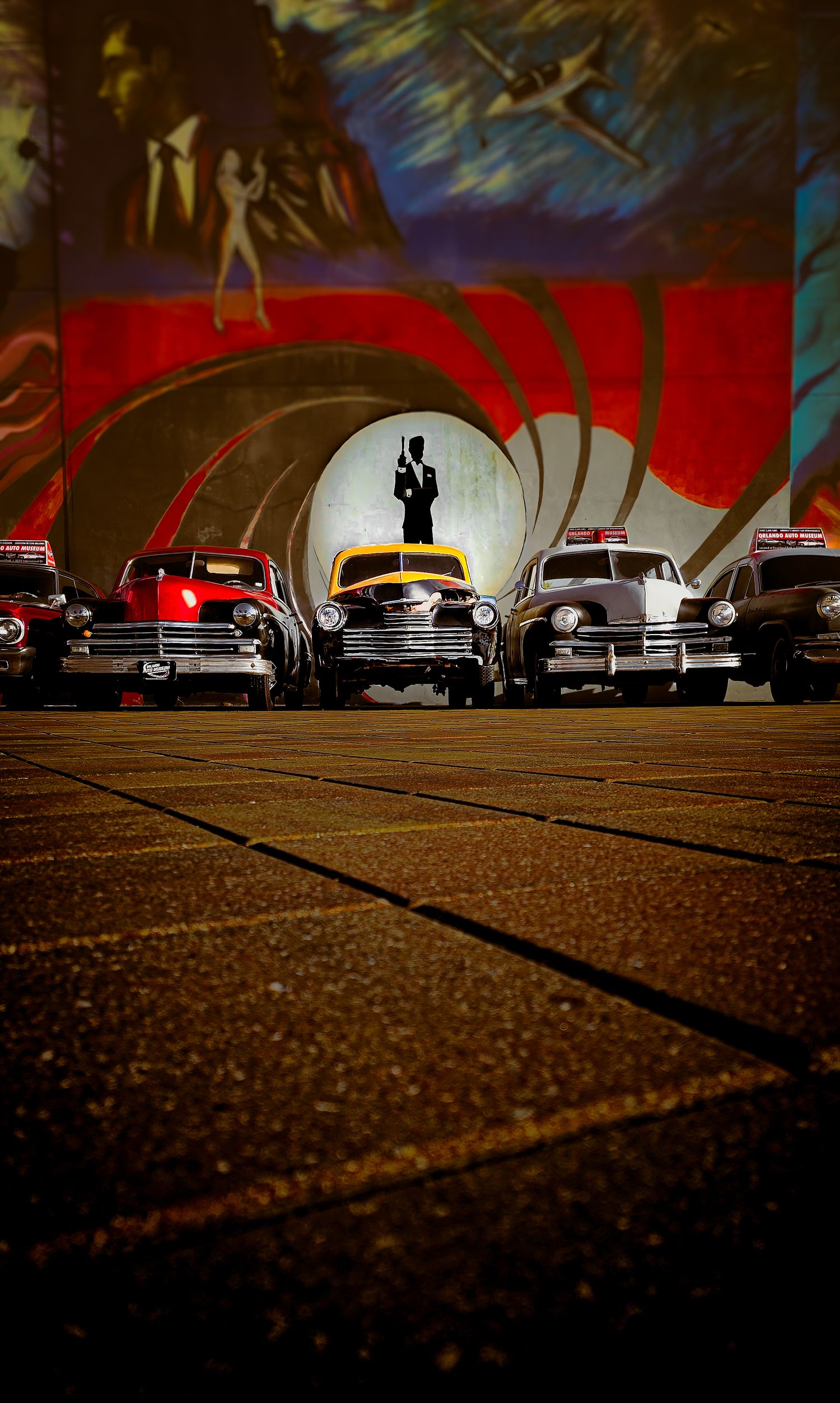 Vintage toy cars lined up in front of a colorful James Bond-themed mural featuring the iconic gun barrel sequence.