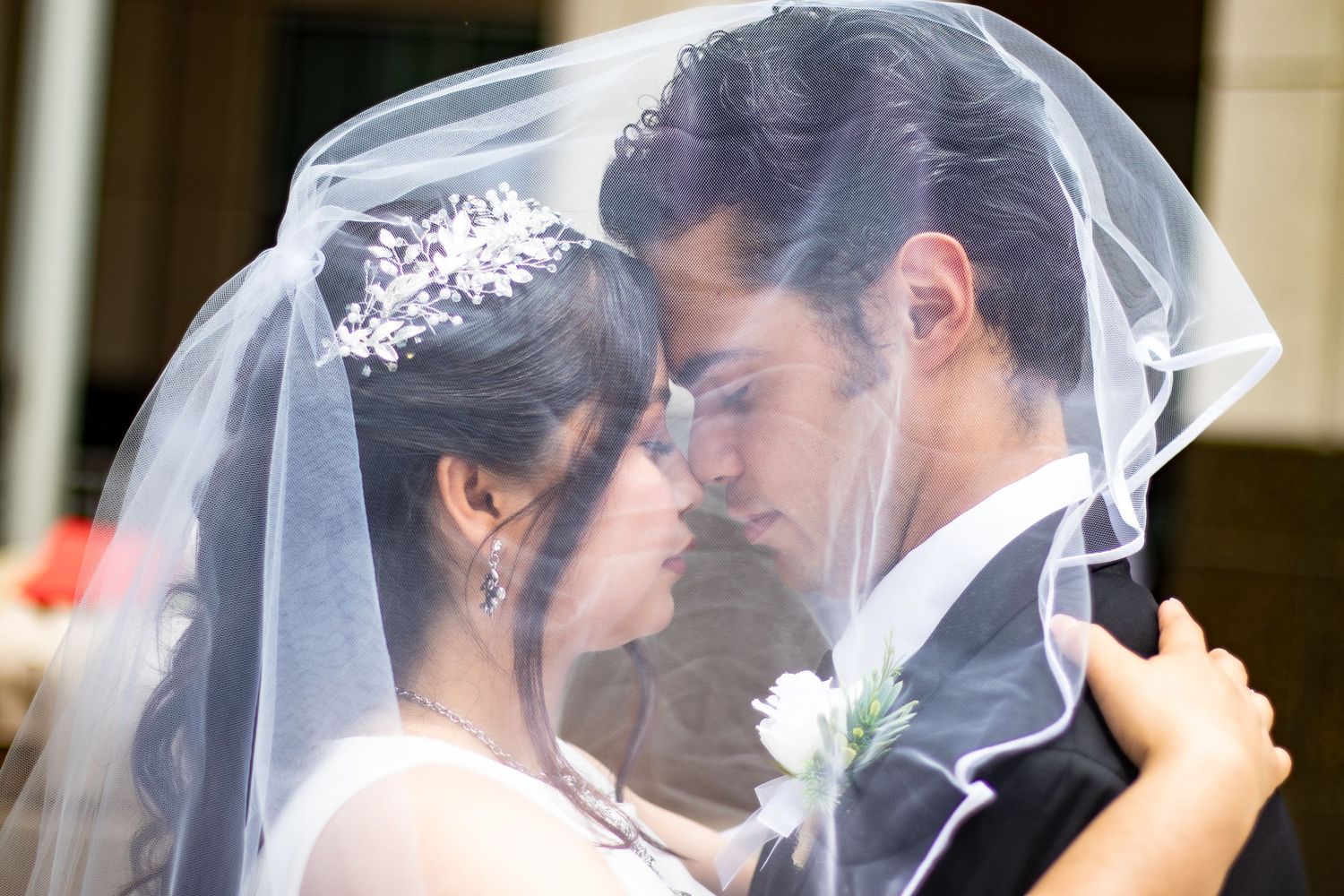 Bride and groom share an intimate moment under a white wedding veil, with the bride wearing a floral tiara.