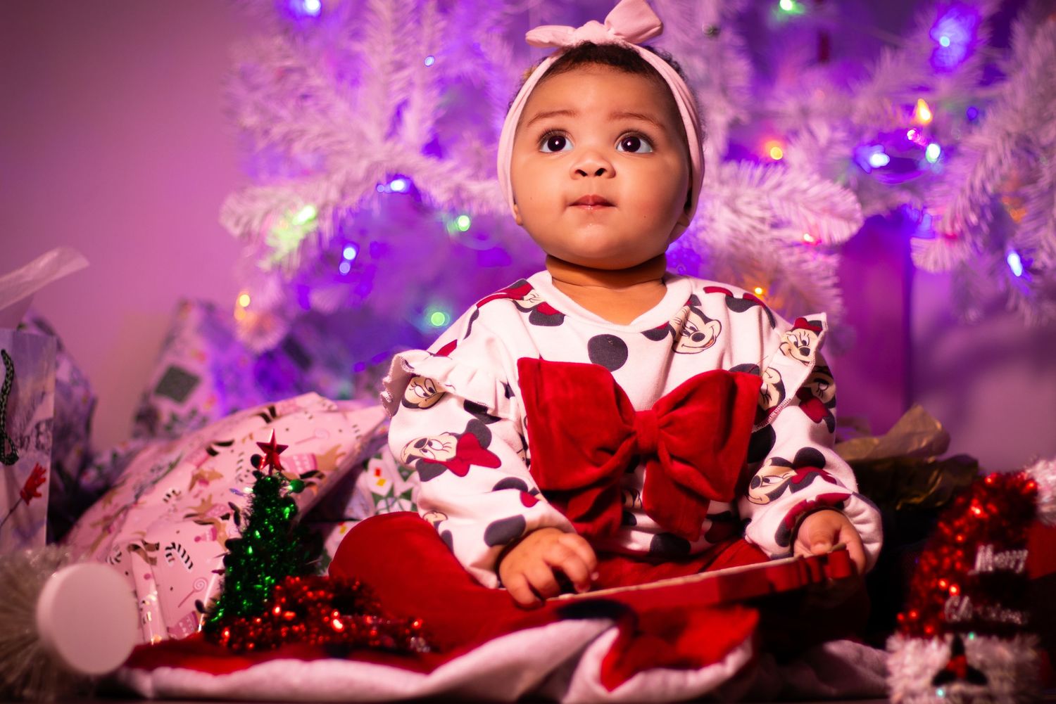 Baby in red holiday outfit poses against purple lit background with Christmas decorations.