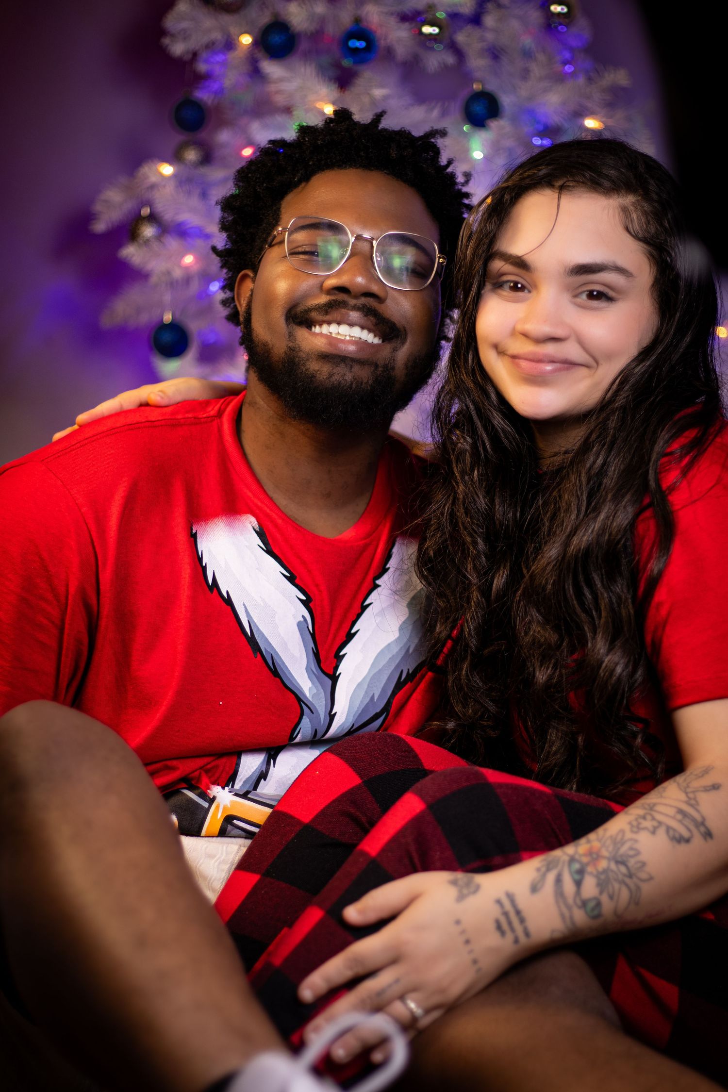 Couple in red shirts share intimate moment under purple holiday lights.