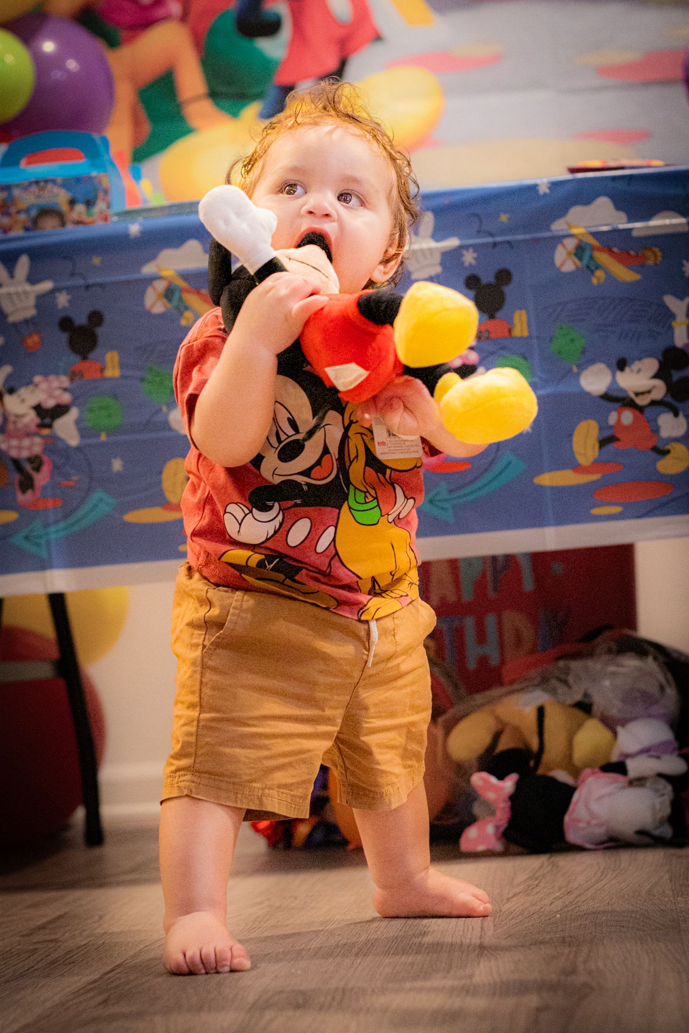 A toddler in khaki shorts and Mickey Mouse shirt plays with colorful toys against a Disney-themed wall border.