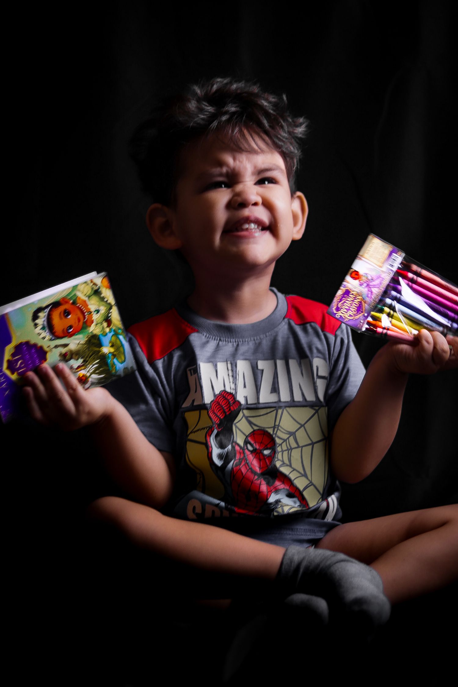 Young boy in Spider-Man shirt smiling while holding coloring book and colored pencils against dark background.