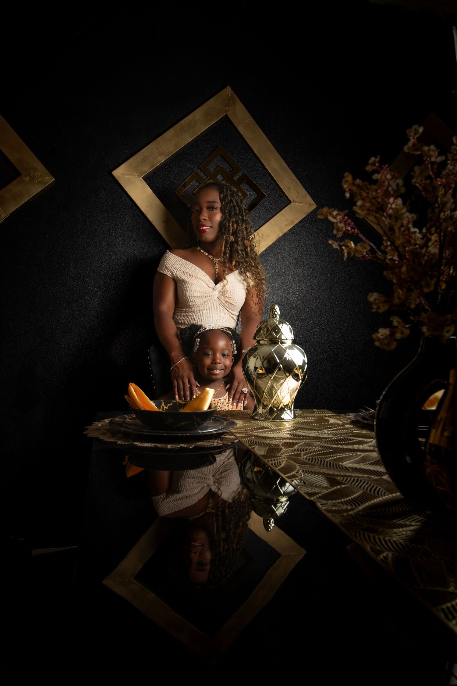 A woman and child pose elegantly at a decorated table with gold accents against a dark backdrop.
