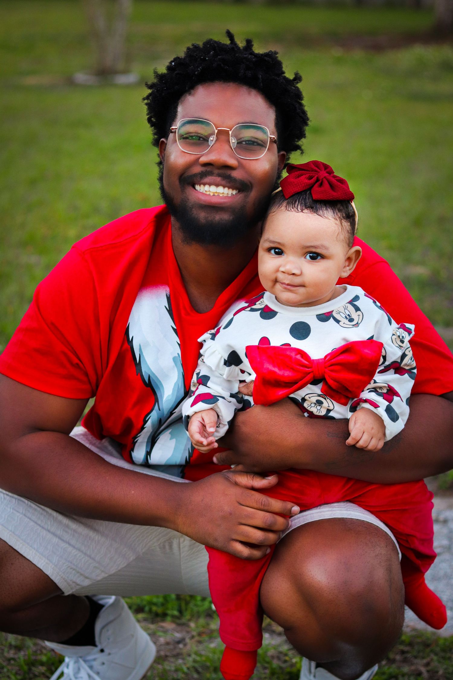 Family member in red shirt holds smiling baby wearing Mickey Mouse outfit on grass.