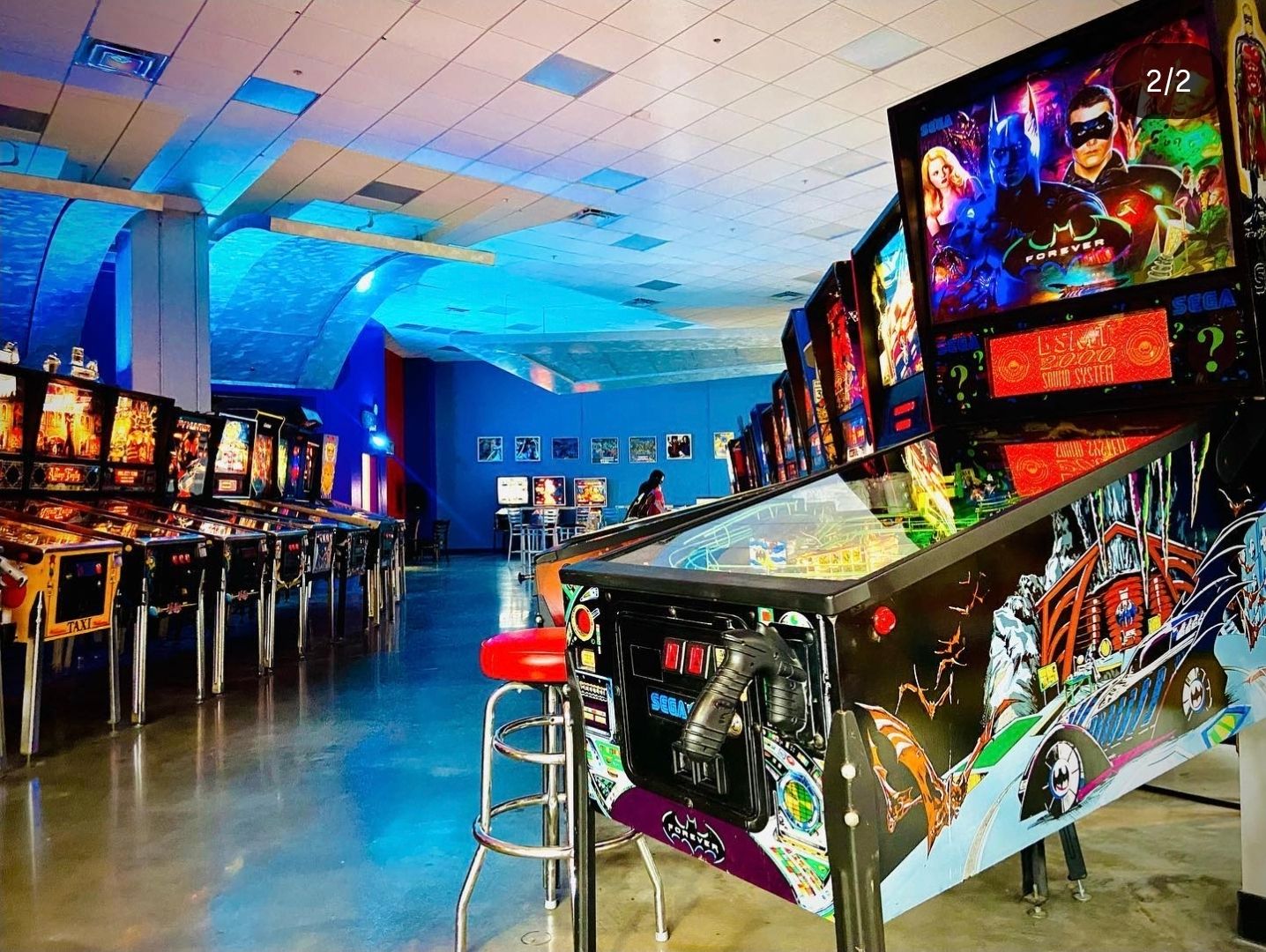 Arcade room with pinball machines illuminated by blue lighting and neon signs.