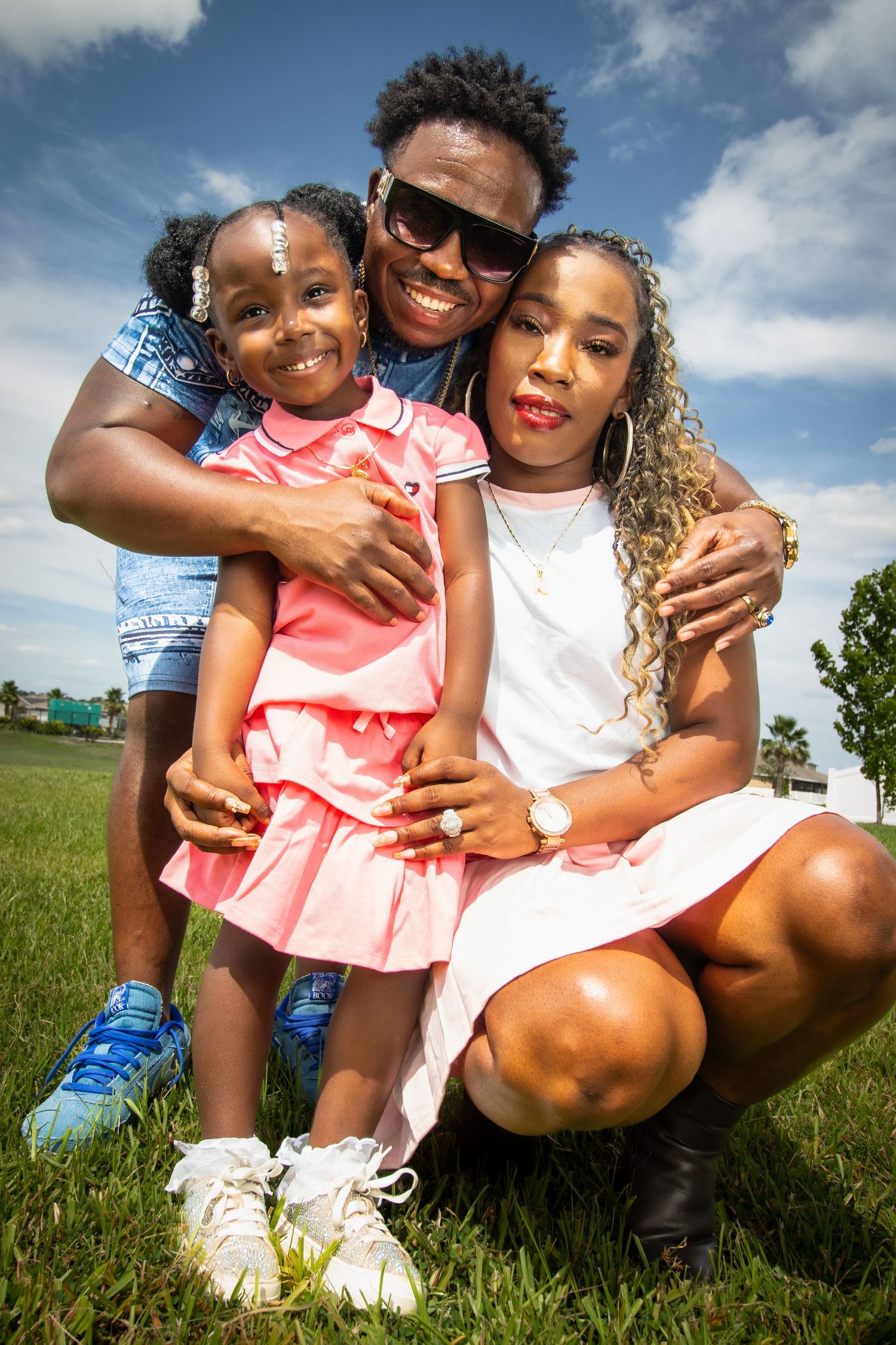 Happy Black family of three posing outdoors on green grass under blue sky, father hugging daughter and mother together.
