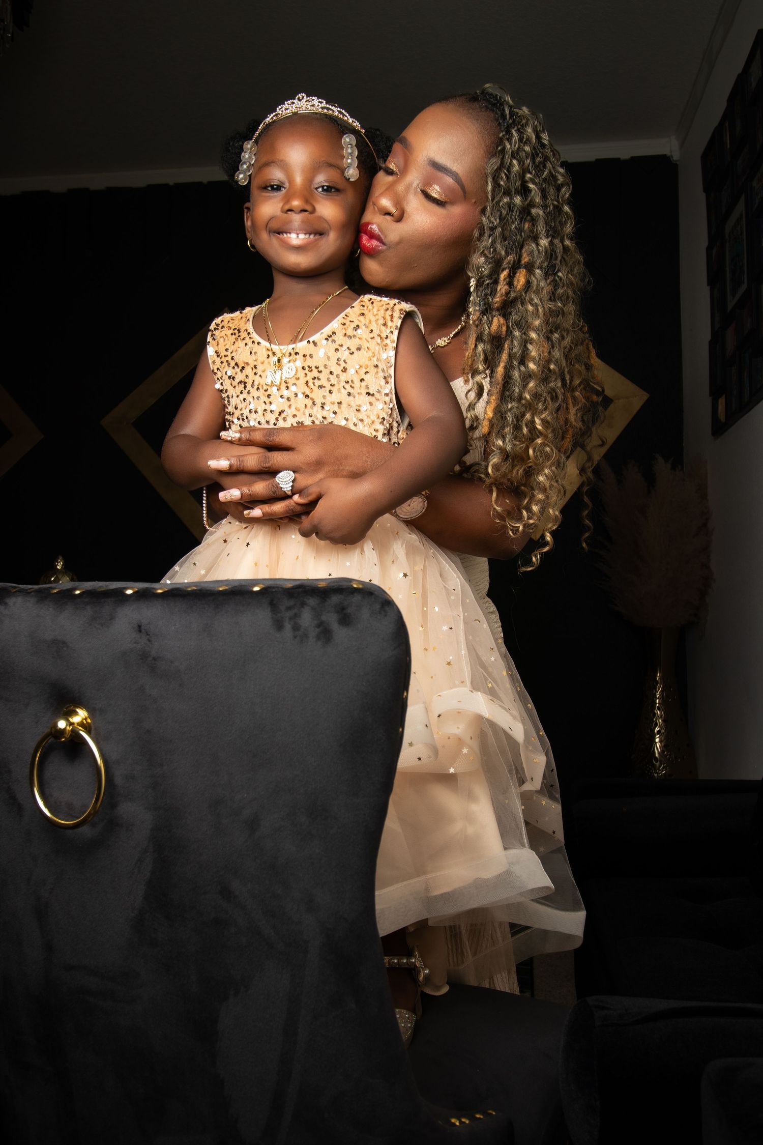 A woman with curly hair kisses a young girl in a tiara and gold dress, both dressed elegantly in a dark room.