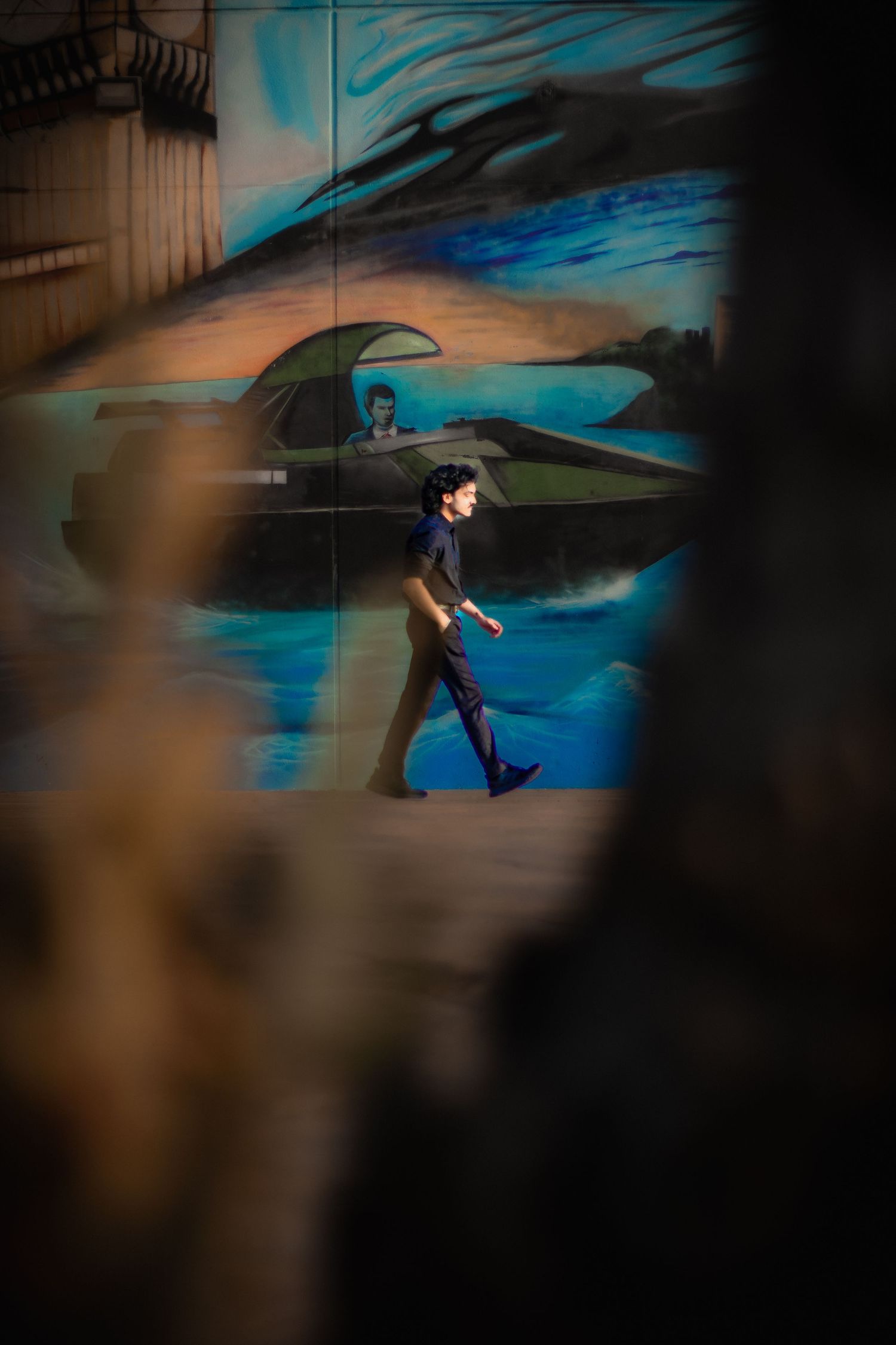 A person walks past a vibrant blue street mural featuring a futuristic vehicle, framed by blurred foreground elements.