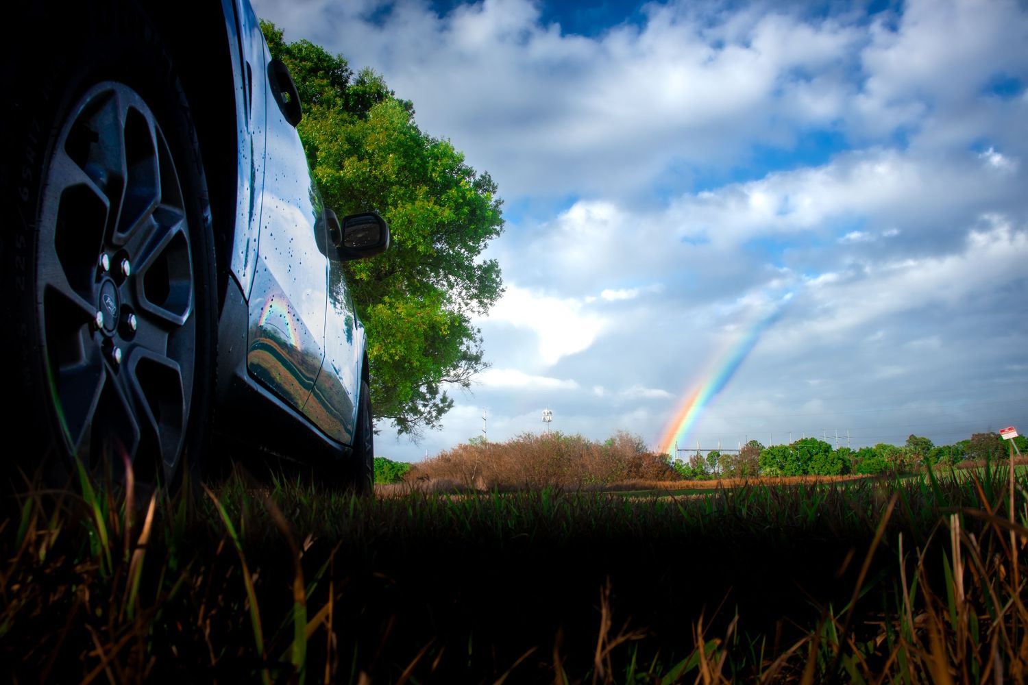 Low-angle view of a car wheel beside a grassy field with a vibrant rainbow and lush green trees under a cloudy sky.
