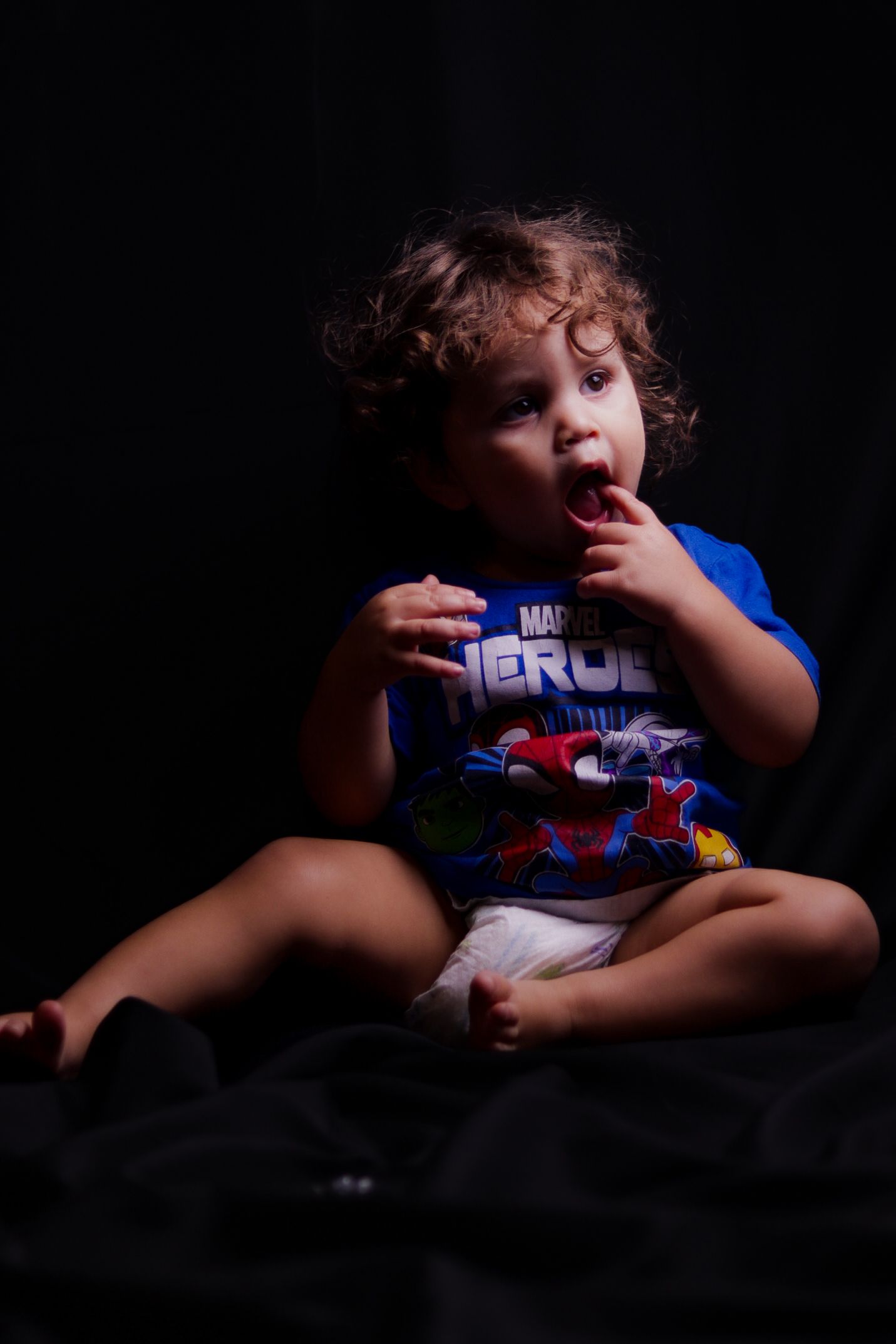 Toddler with curly hair wearing a Marvel Heroes blue shirt sits on dark background, fingers near mouth.