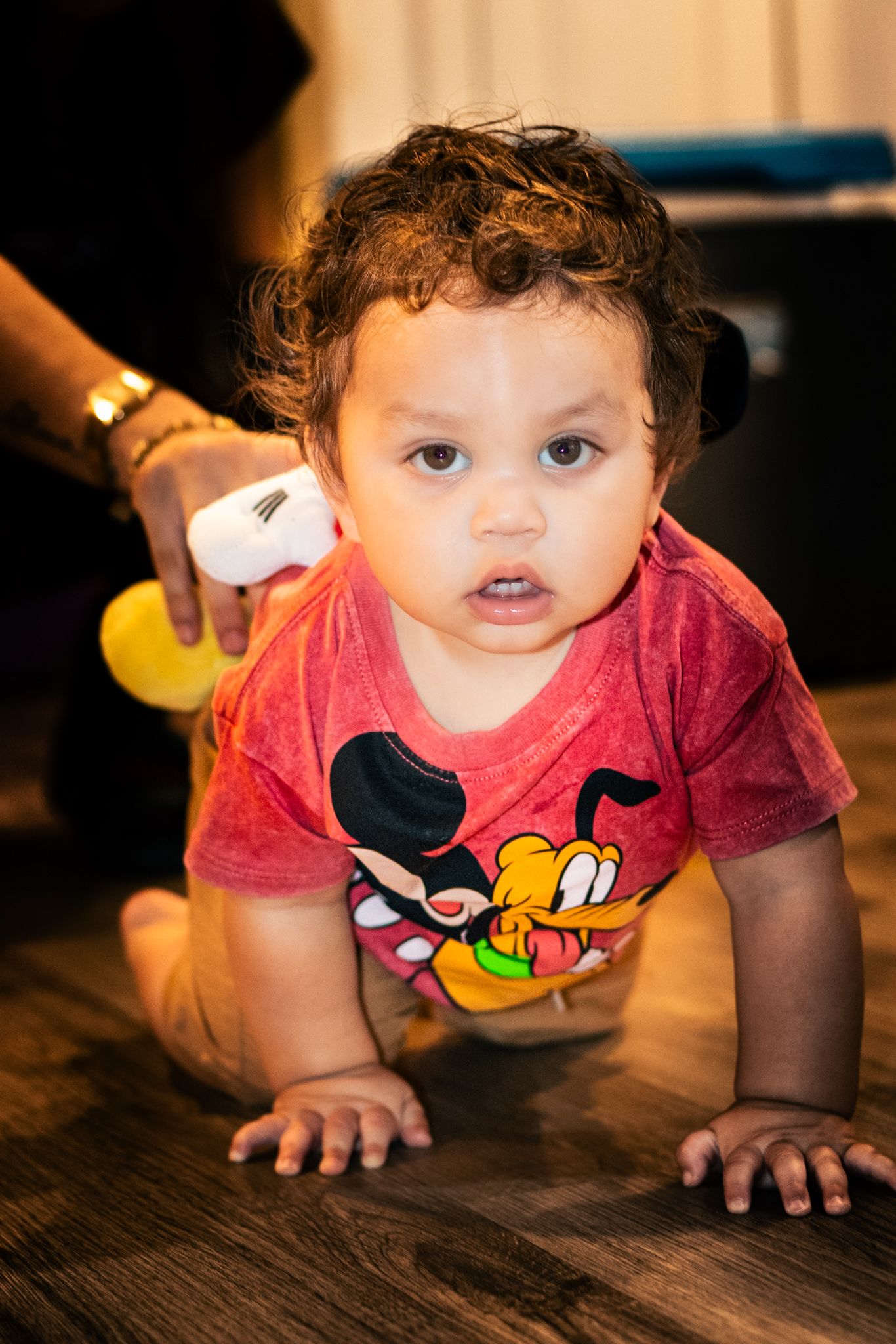 A small child crawls on hardwood floor wearing a red Mickey Mouse t-shirt.