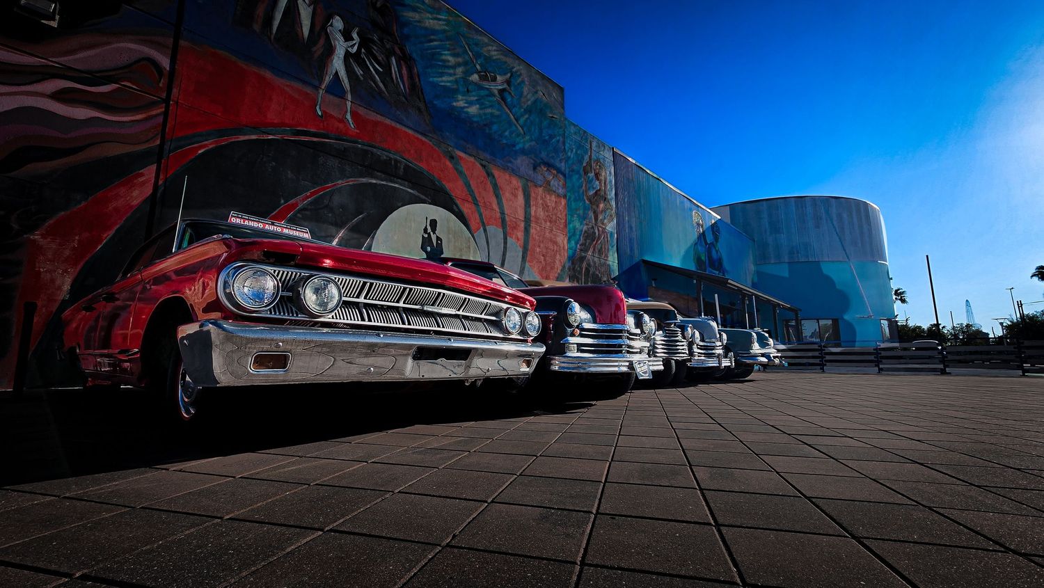 Classic American cars lined up outside a colorful mural building under a bright blue sky.