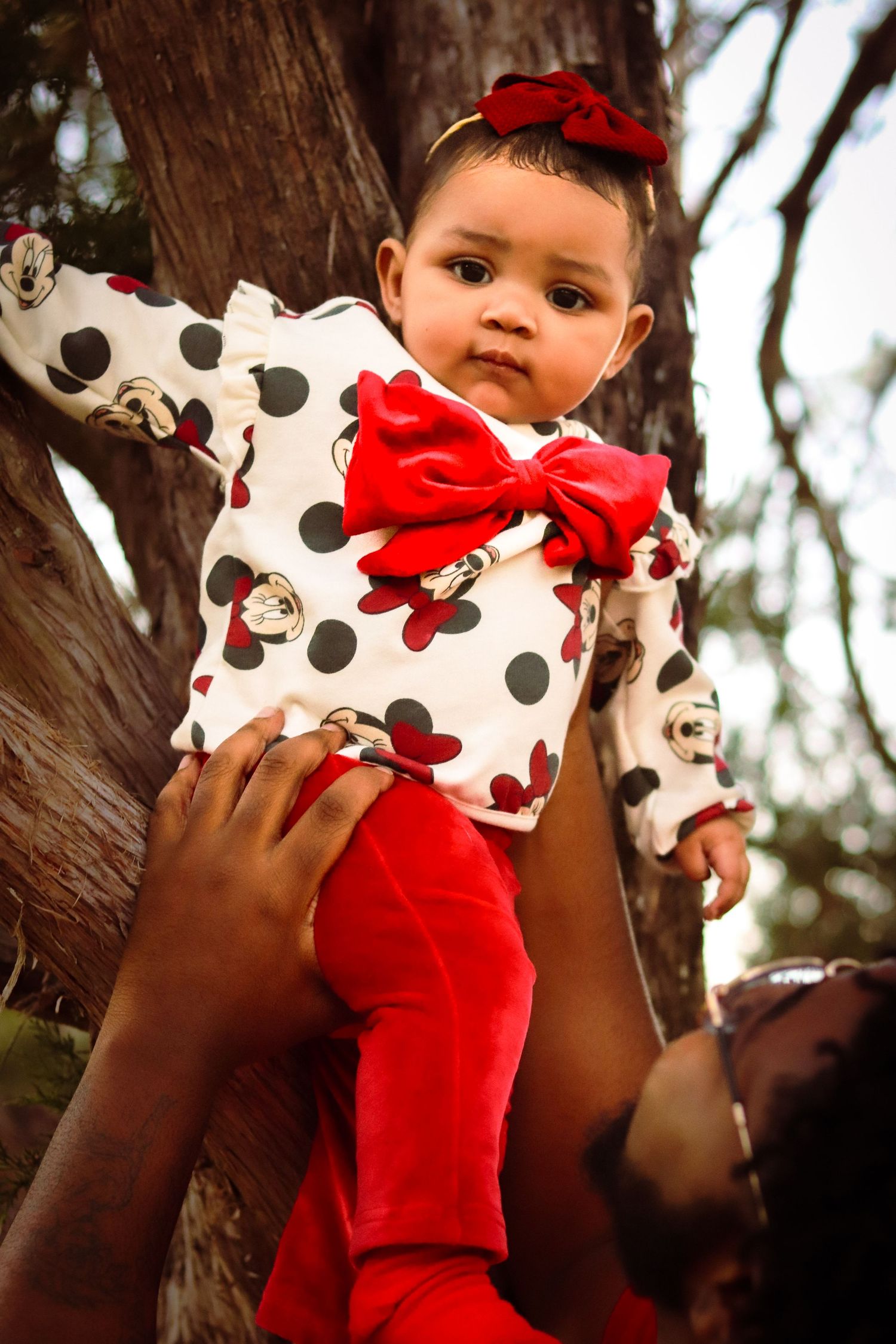 Baby wearing polka dot outfit with red bow being held up against tree backdrop.