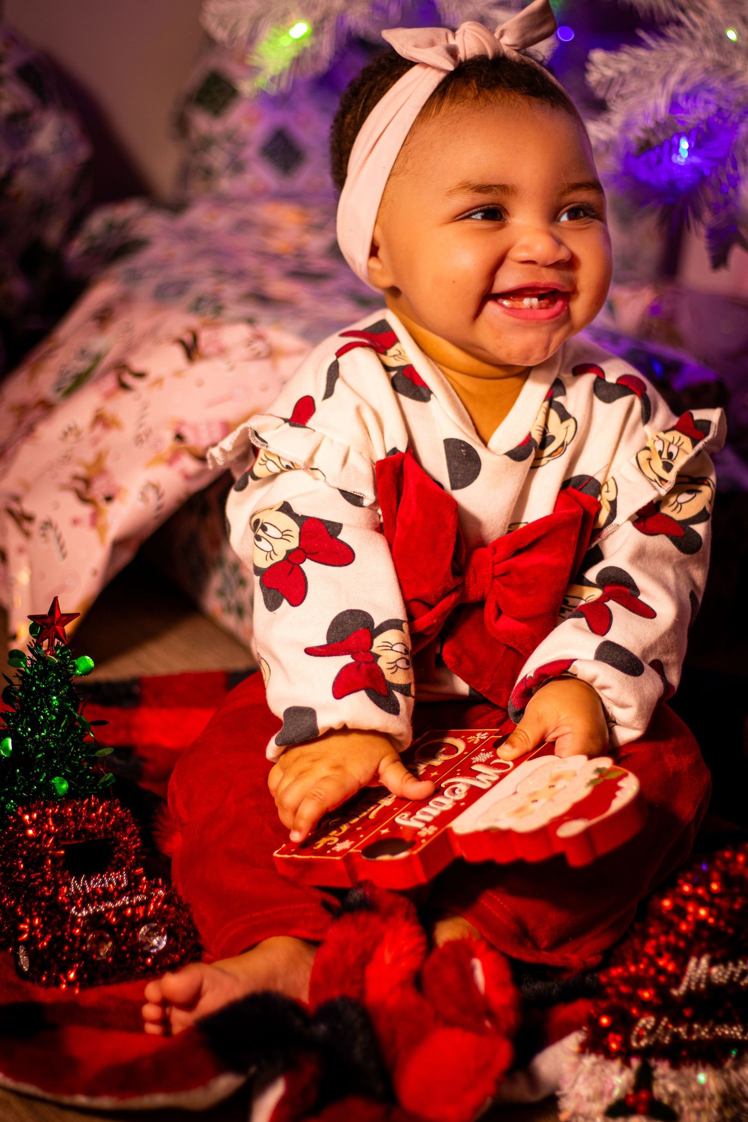 Baby wearing festive holiday outfit sits on red blanket surrounded by Christmas decorations.
