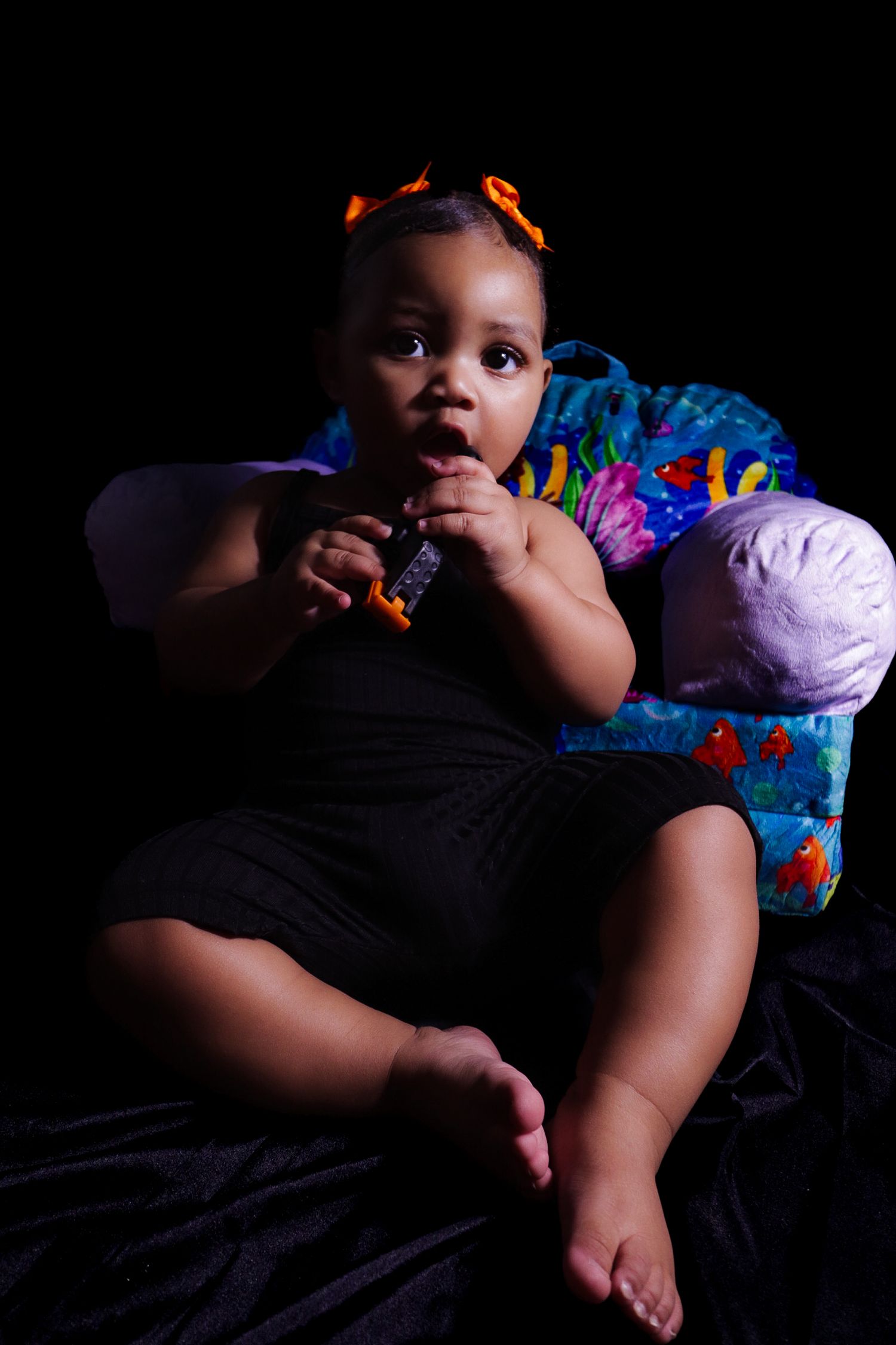 Adorable baby girl with orange hair bows sitting in colorful chair, chewing toy against black background.
