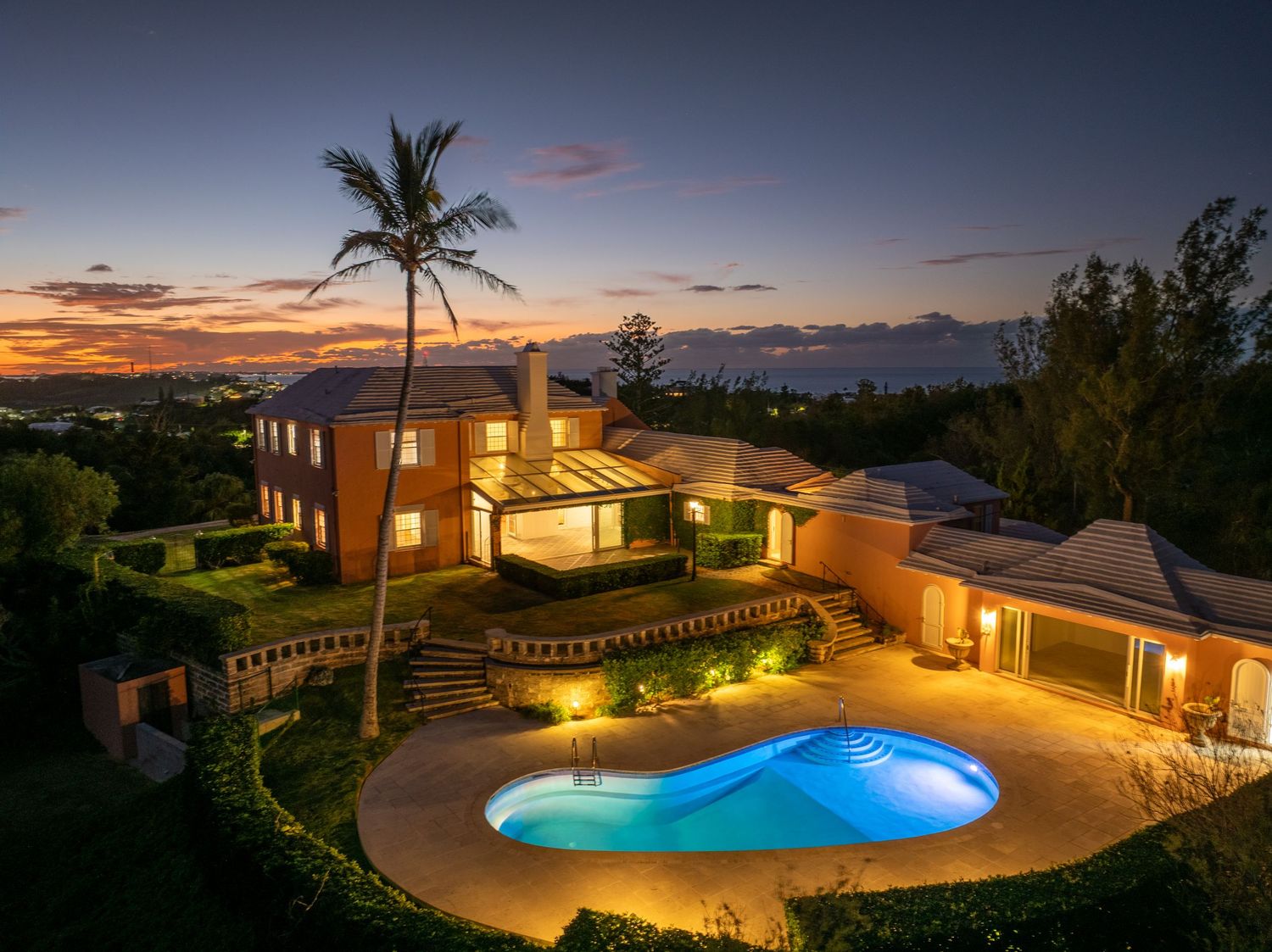 Sunset view of a Mediterranean-style mansion with illuminated pool and ocean vistas. Drone photogrpahy.