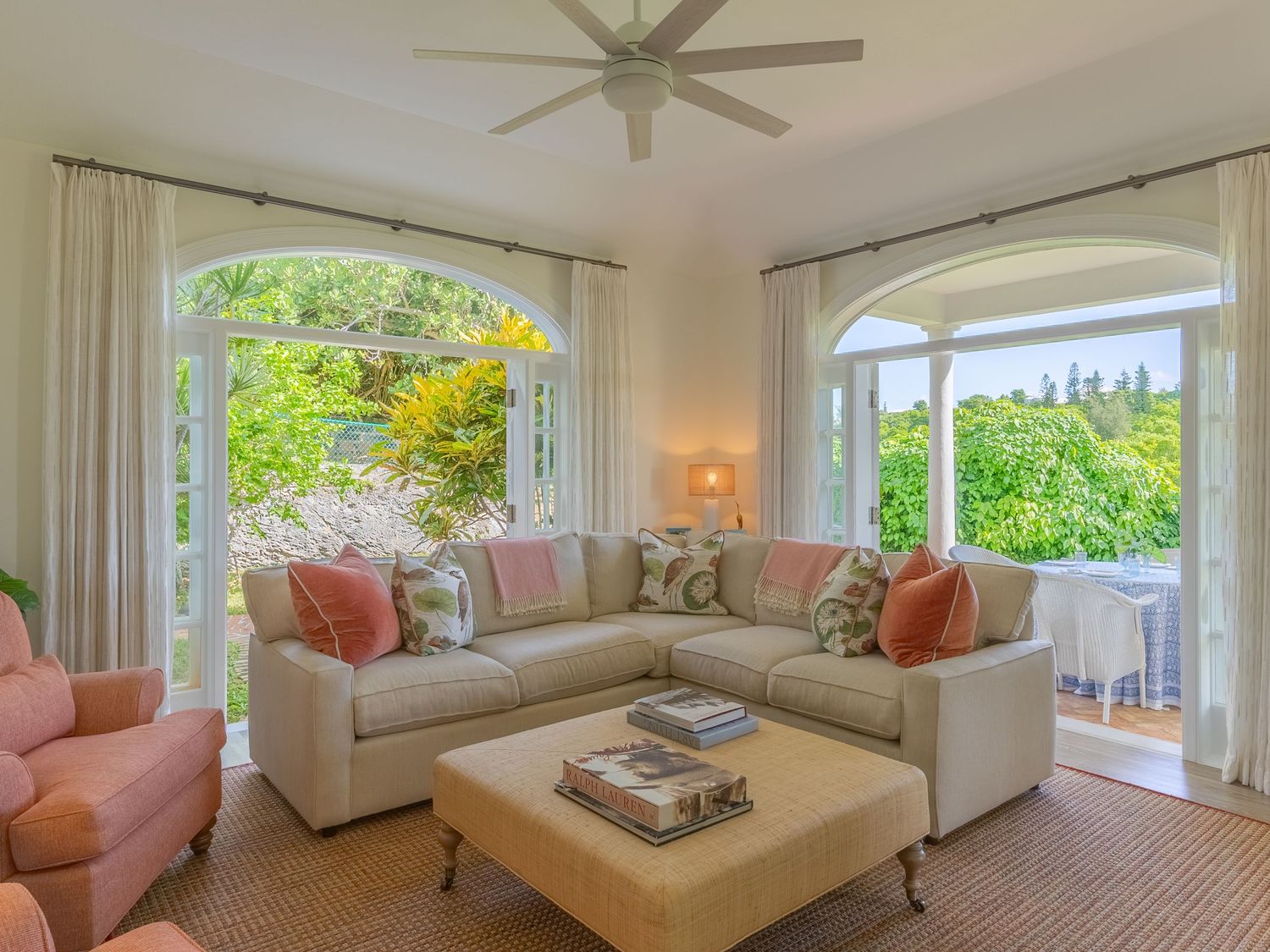 Bright coastal-style living room with ceiling fan, plush seating, and multiple windows offering garden views.